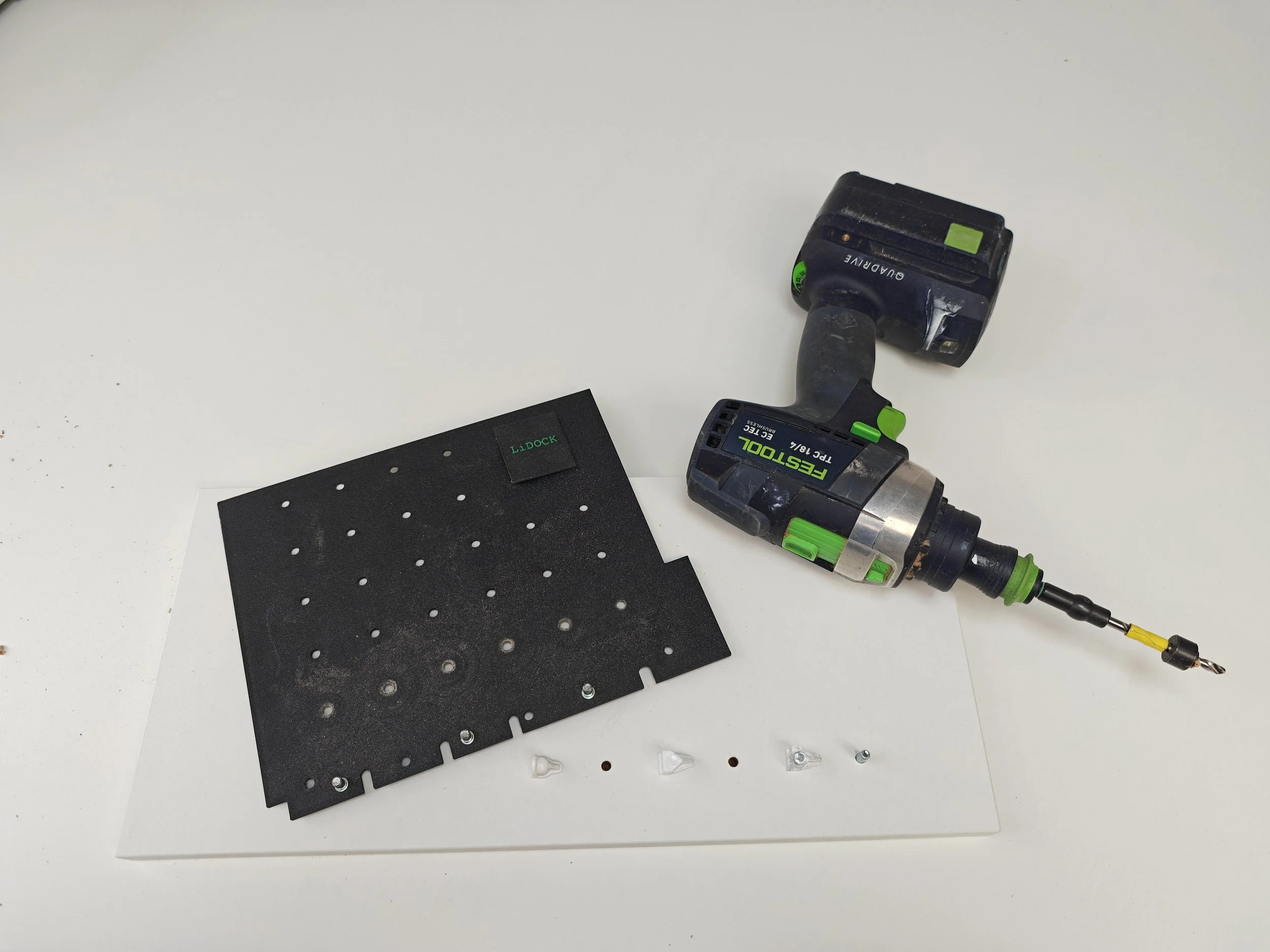 A cordless power drill next to a black perforated cutting board on a white surface, with small plastic and metal hardware components nearby.