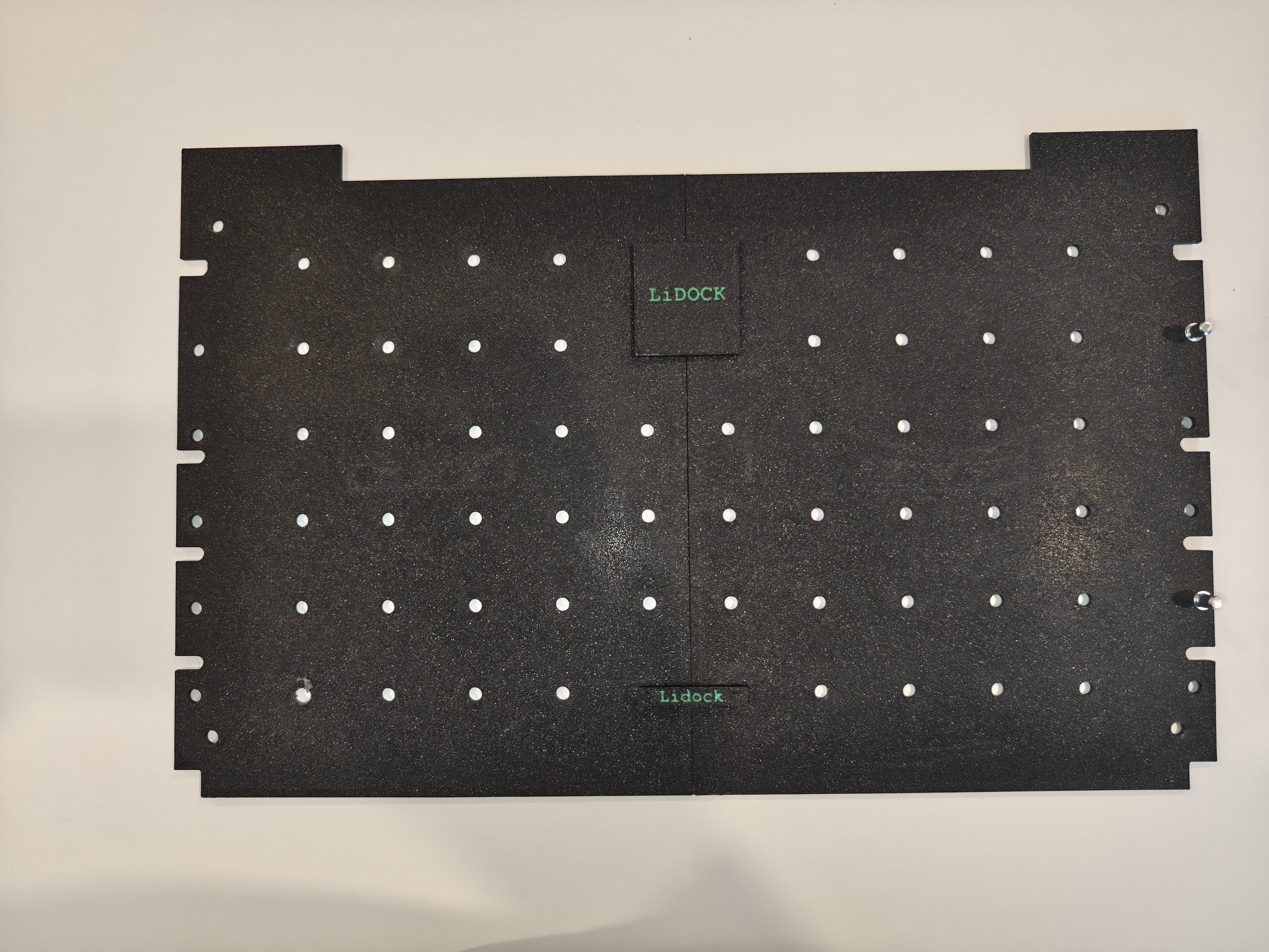A black perforated metal platform with multiple holes, labeled 'LIDOCK' in green, resting on a white surface.