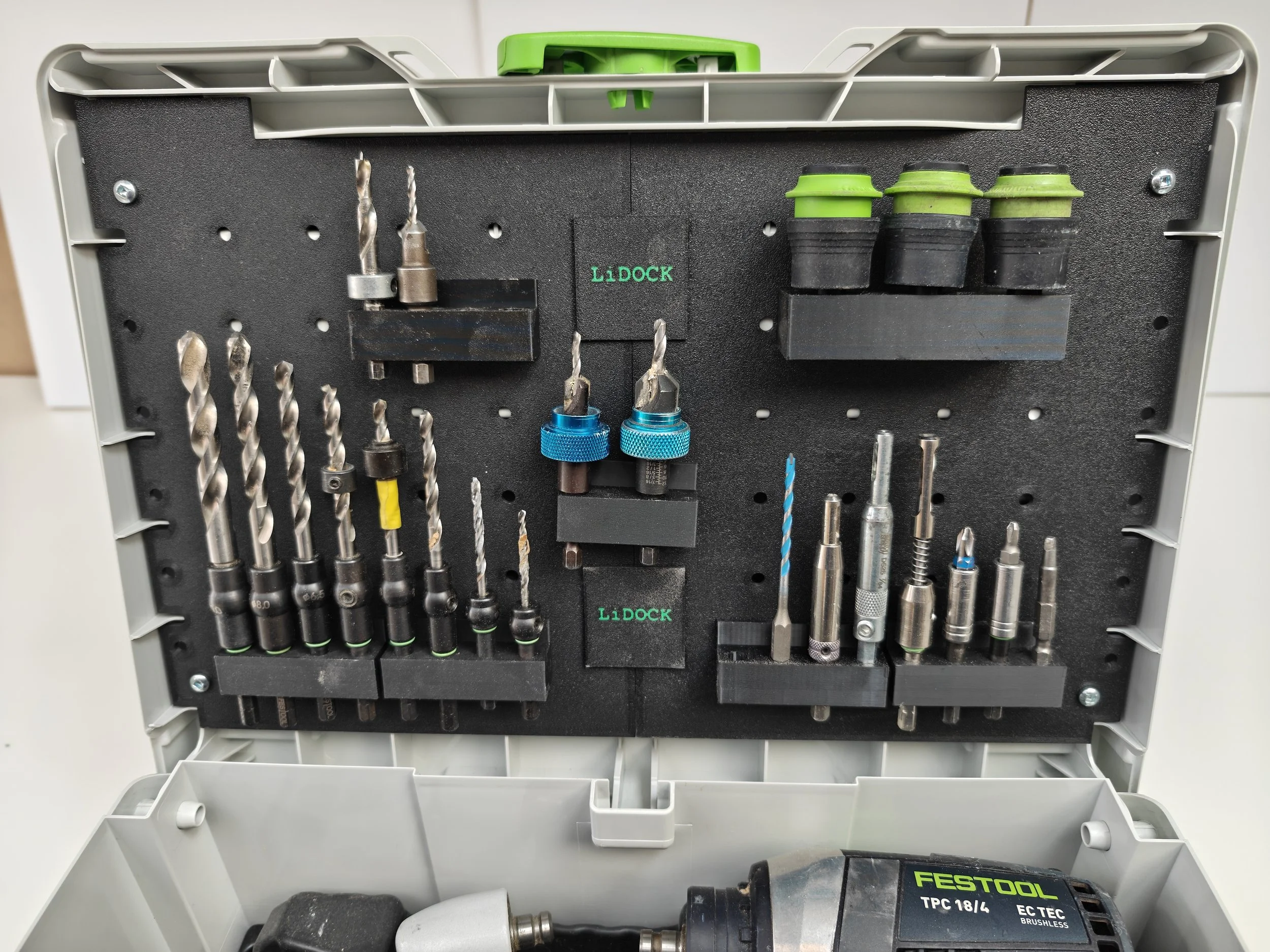 An open plastic tool storage box holding various drill and screwdriver bits, with a green and black drill at the bottom.