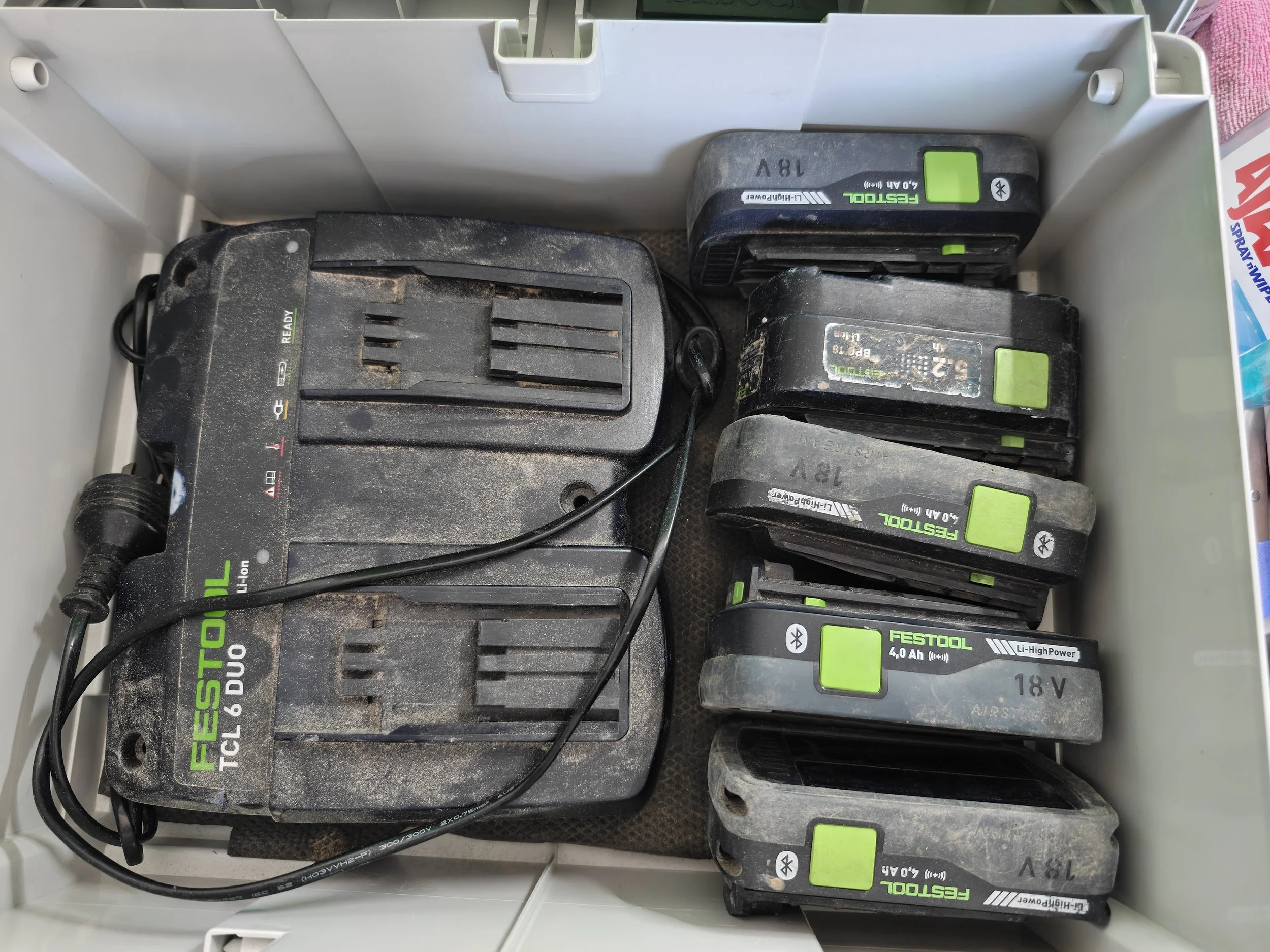 Several power tool batteries stored in a white plastic container. The batteries show signs of dust and wear, with green labels and black casing.