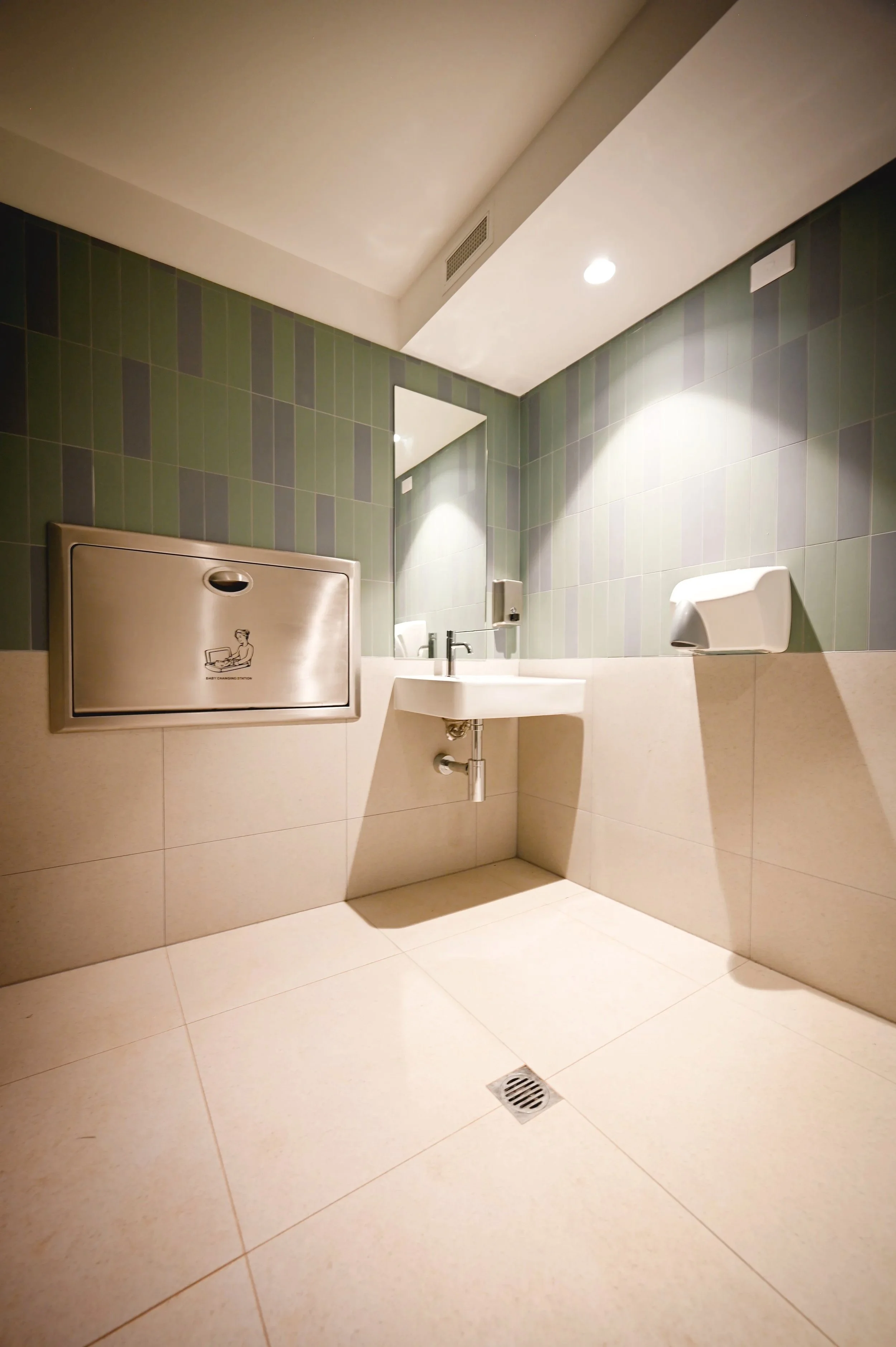 Public restroom with green tiled walls, a mirror, sink, and paper towel dispenser, with a drain on the beige tiled floor.