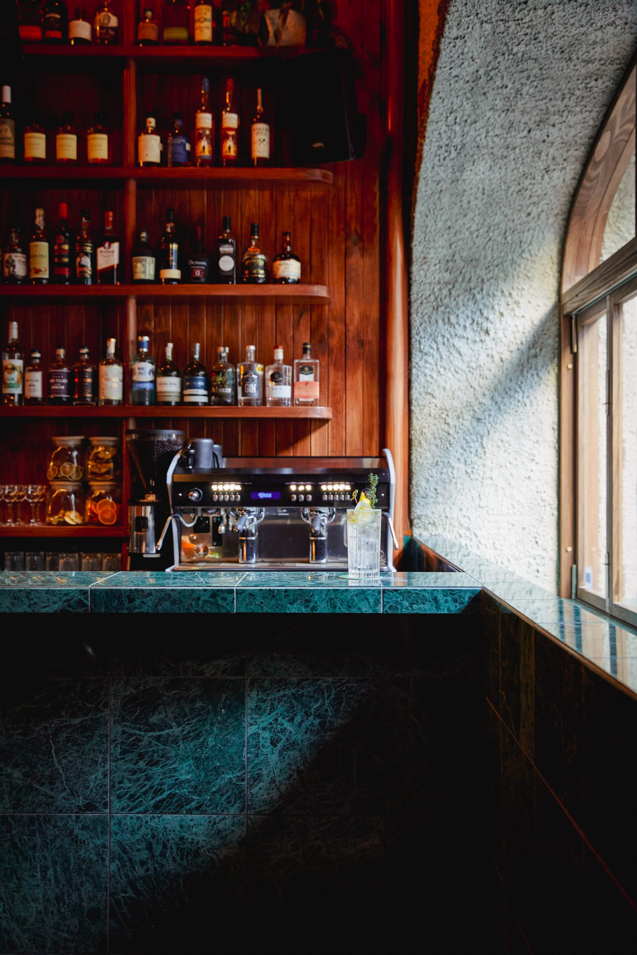 Bar with dark green marble counter, espresso machine, and a glass with a lemon slice, against a wood-paneled wall filled with liquor bottles and a window with natural light.