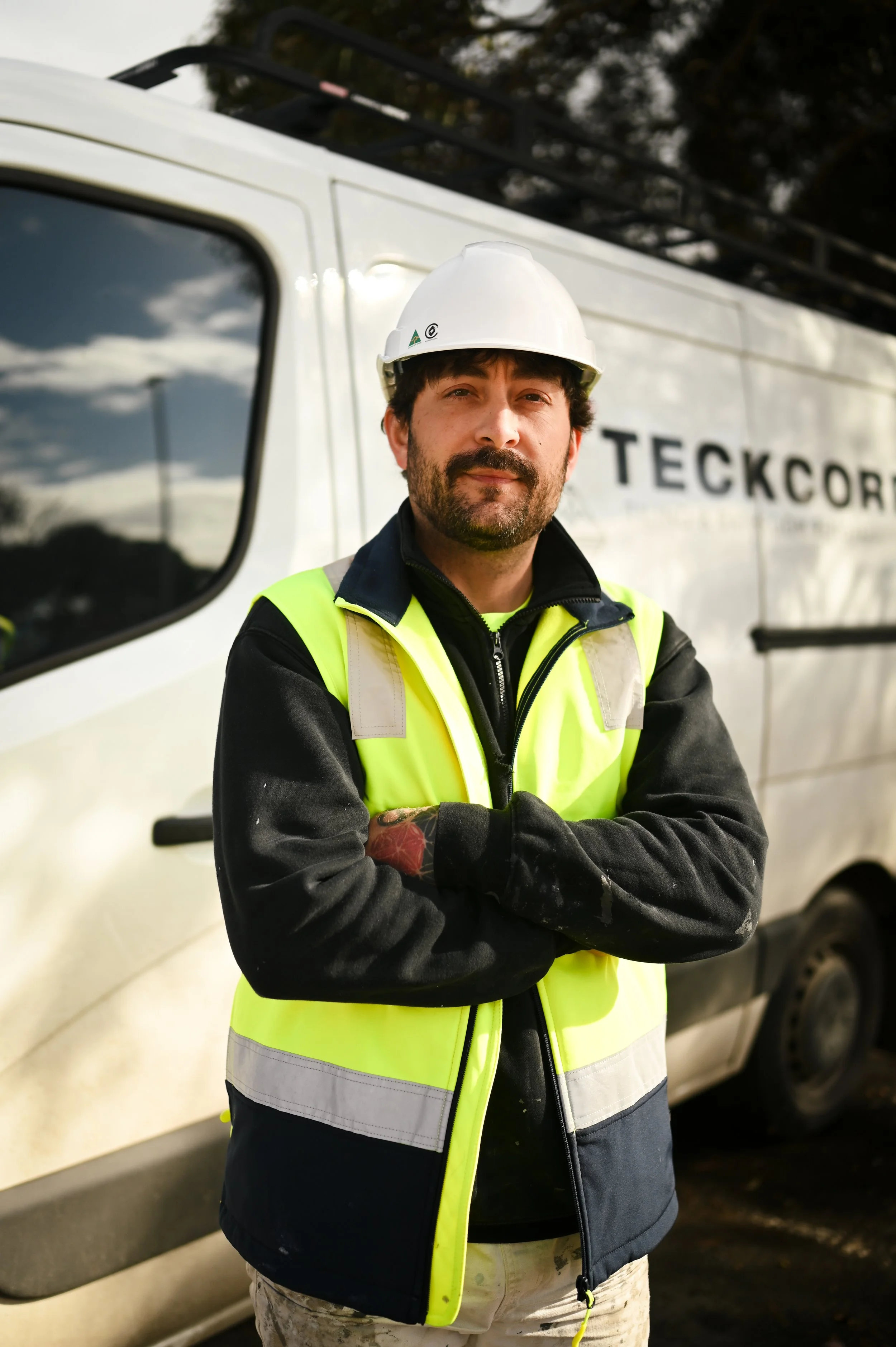 A man wearing a white safety helmet and a high-visibility yellow vest with reflective stripes stands with crossed arms in front of a white work truck. The truck has the word 'TECKCOR' partially visible on its side. The man has dark hair, a beard, and mustache, and is wearing a black jacket underneath the vest. The background shows trees and the reflection of clouds on the truck window.