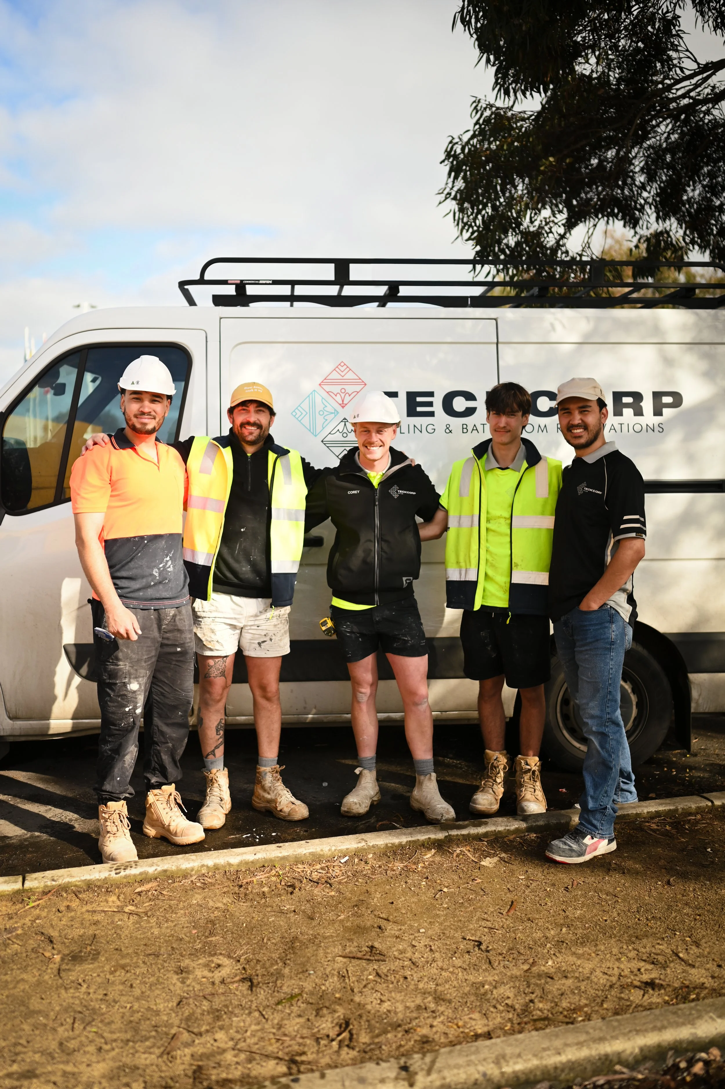 A group of five construction workers standing in front of a white van with company branding, wearing safety helmets and reflective vests, smiling.