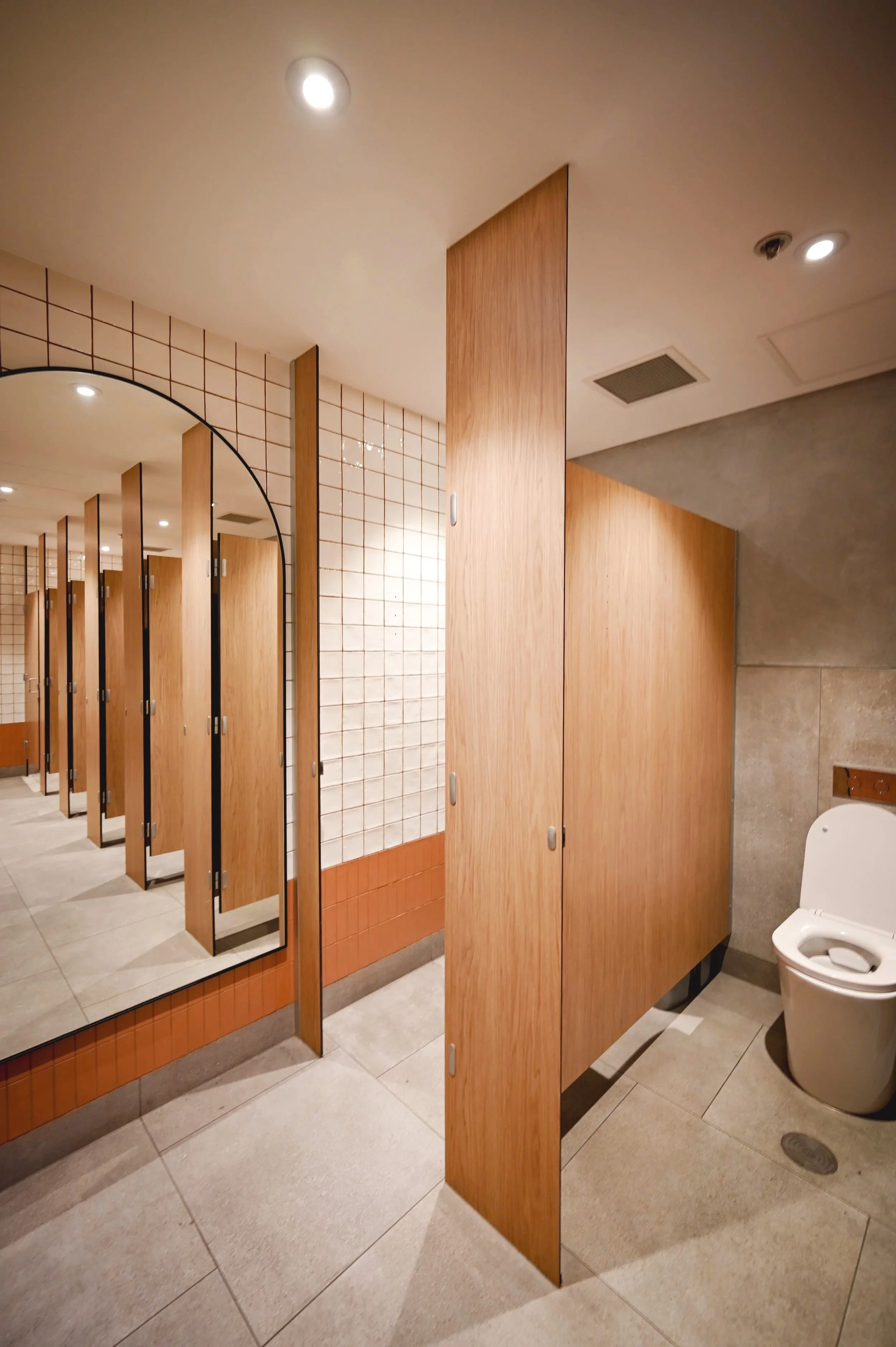 Men's restroom with wooden stalls, a large mirror, beige tile walls, and a toilet. Overhead lighting and a floor drain are visible.