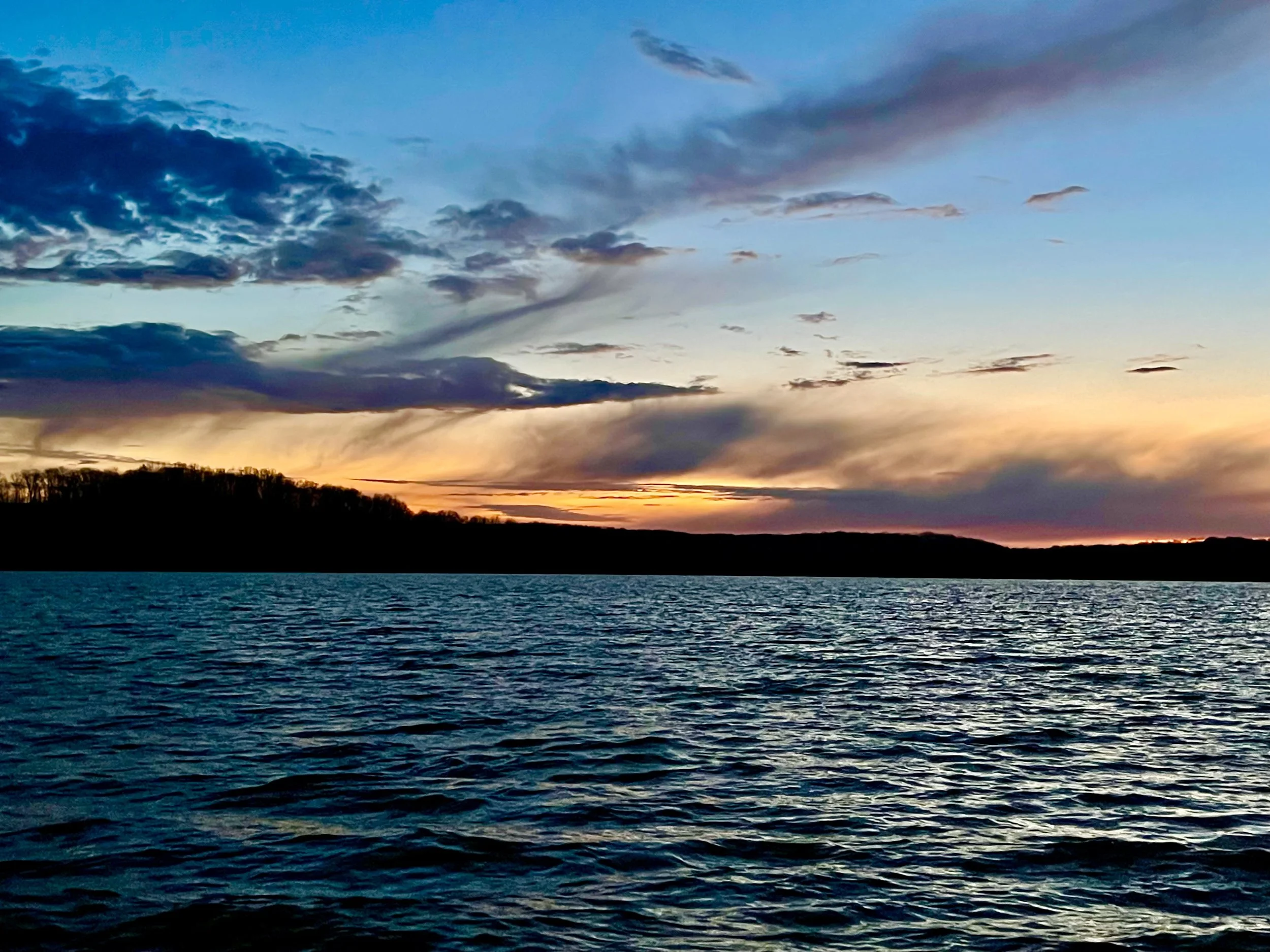 Sunset over a body of water with a silhouette of a landmass or forested area on the horizon, partly cloudy sky, and a calm water surface.