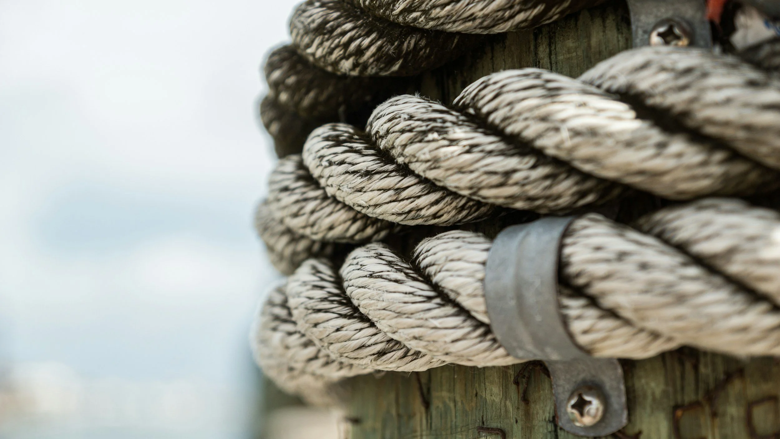Close-up of weathered beige and dark brown twisted nautical rope tied around a wooden post with metal fixtures, against an overcast sky.