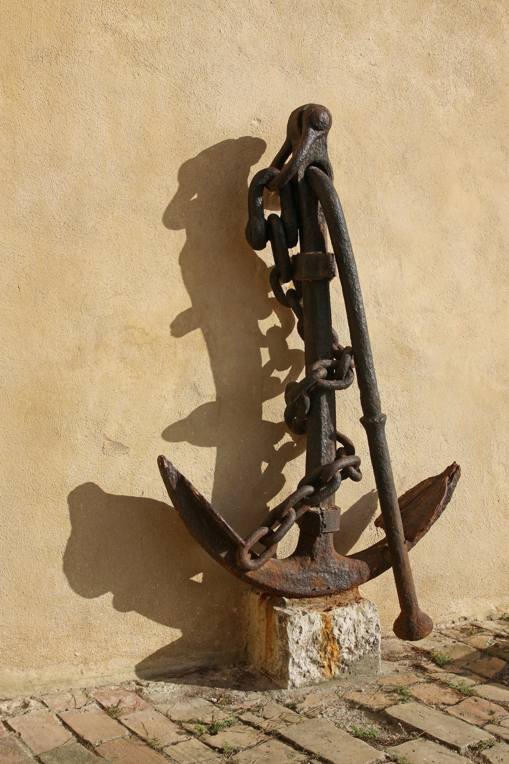 A metal anchor sculpture with a chain against a beige wall, casting a shadow, with a stone base and brick sidewalk at the bottom.