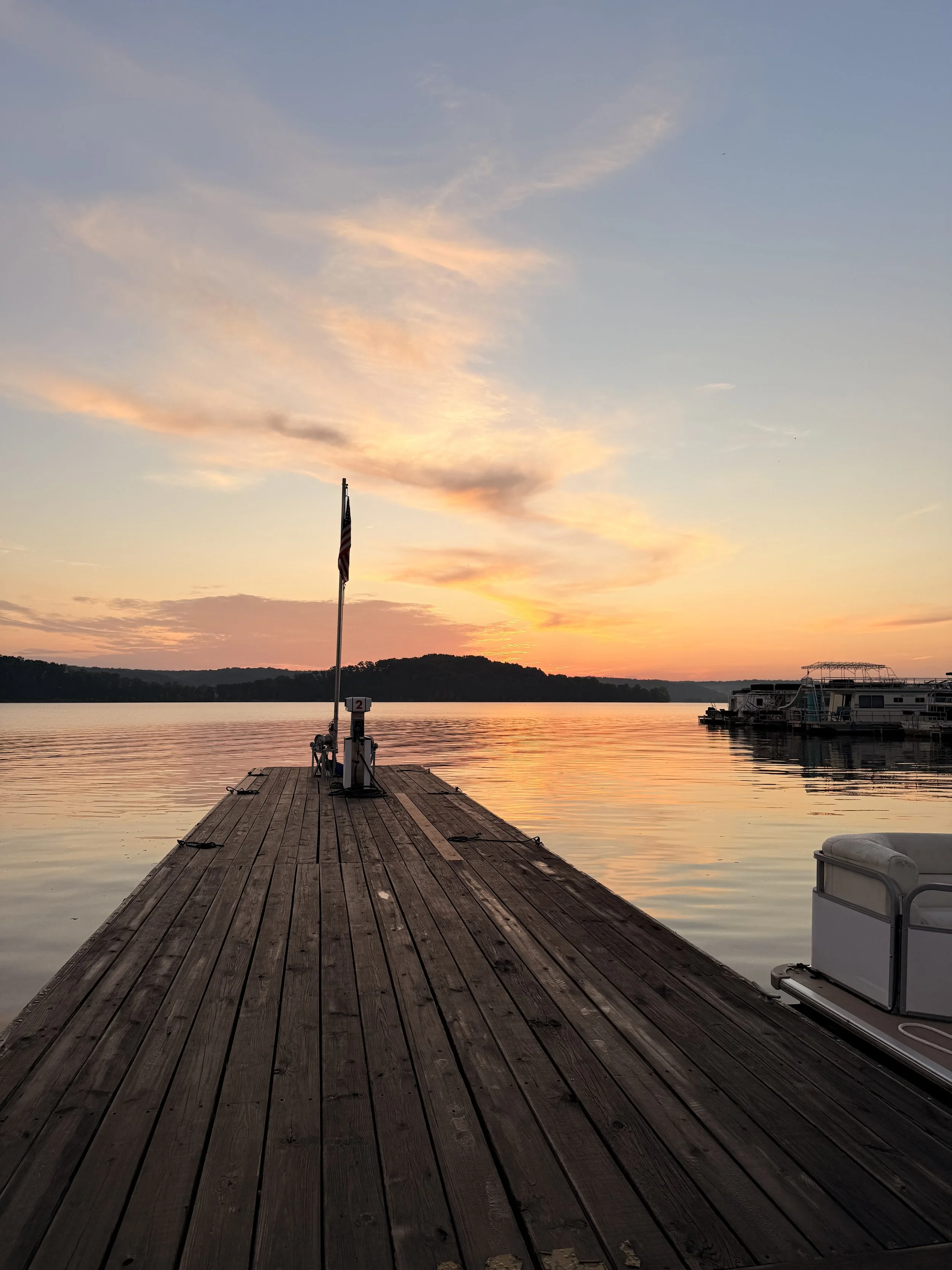 A wooden dock extending into a calm lake during sunset with a flagpole flying an American flag, and boats docked on the right side of the image.