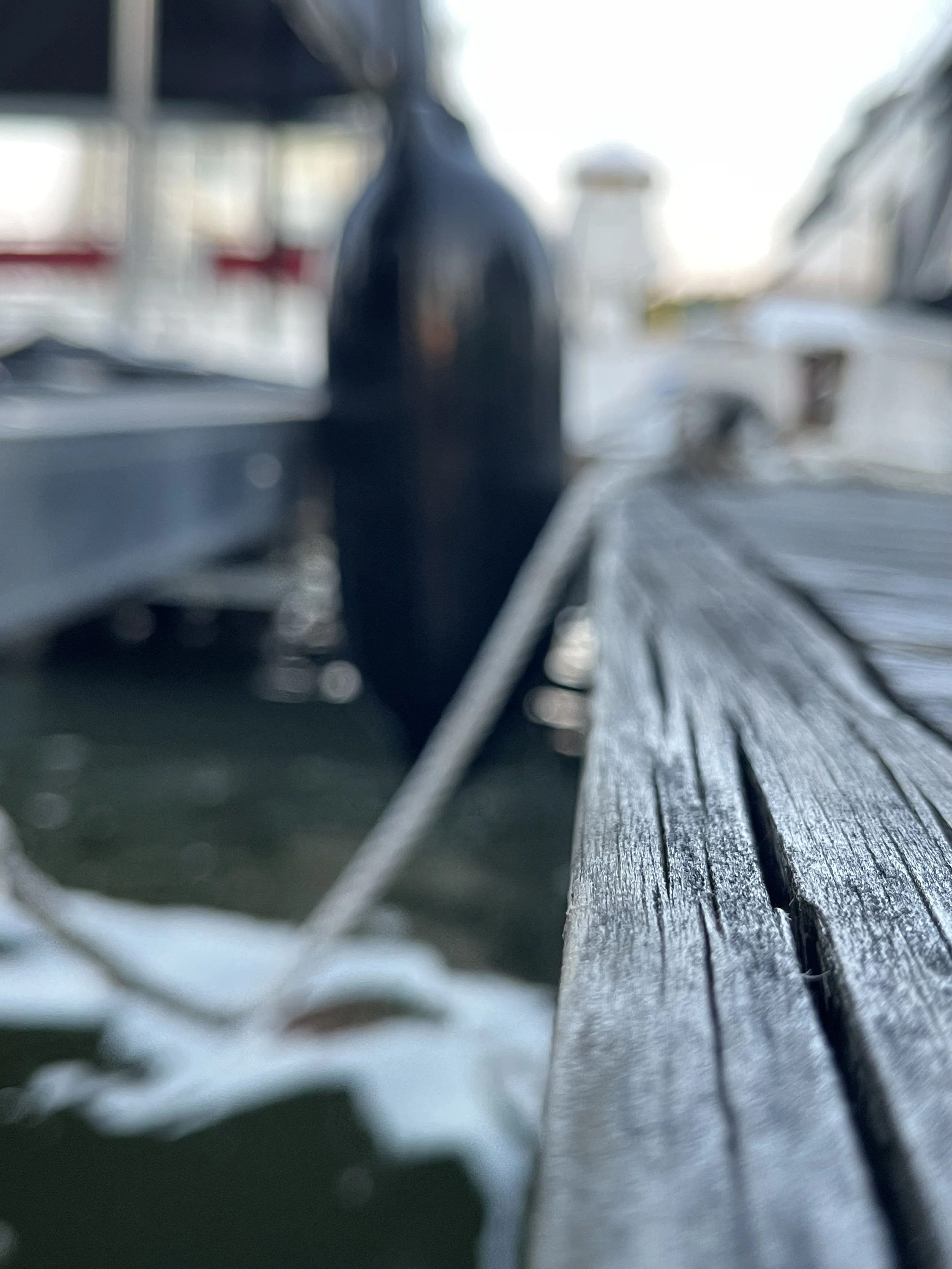 Close-up of a weathered wooden surface, possibly a deck or table, with blurred background that includes a bottle and some objects, in an outdoor setting with bright lighting.