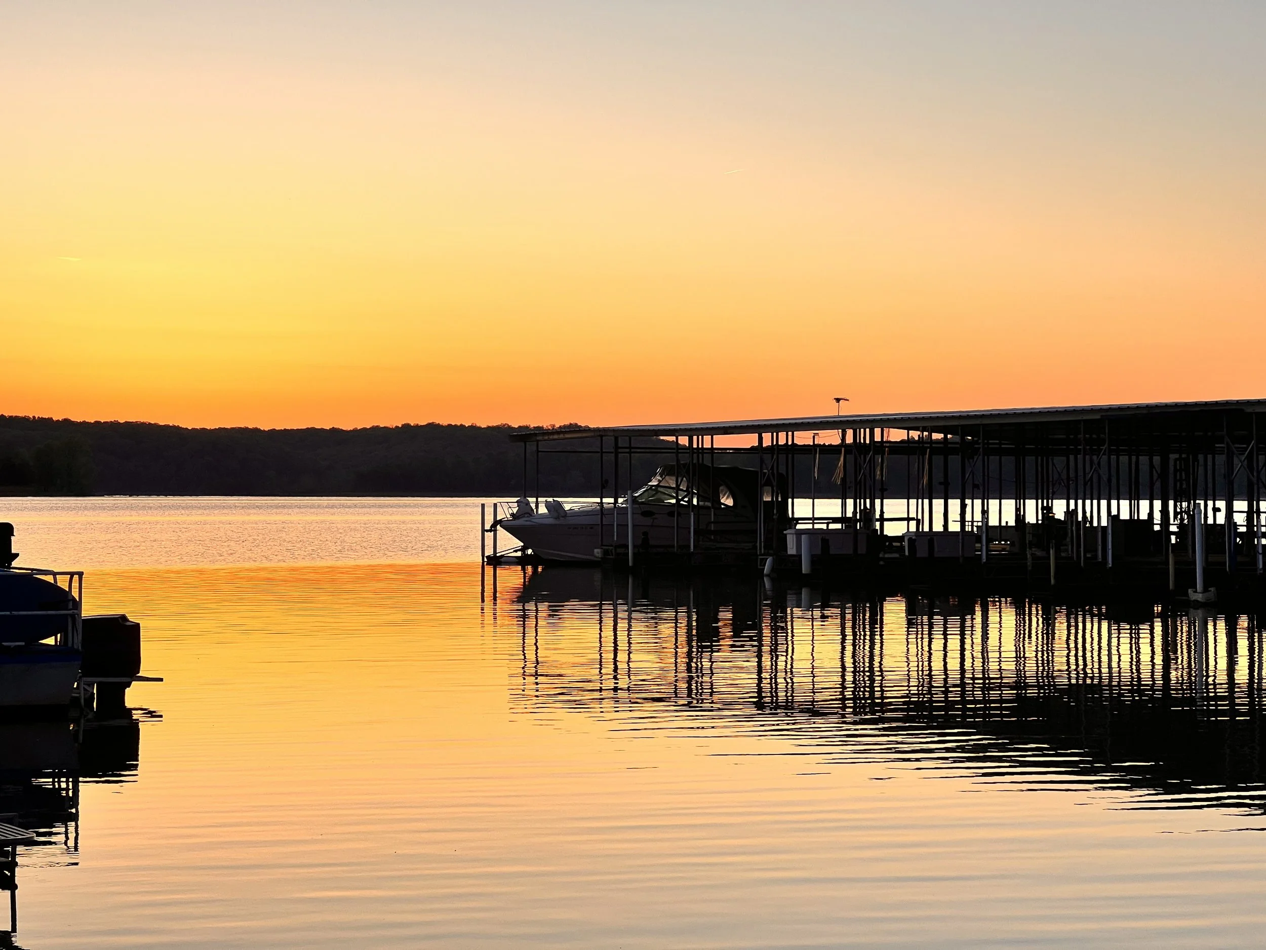 Sunset over a lake with a dock and boats, silhouetted trees in the distance, warm orange sky reflected in the water.