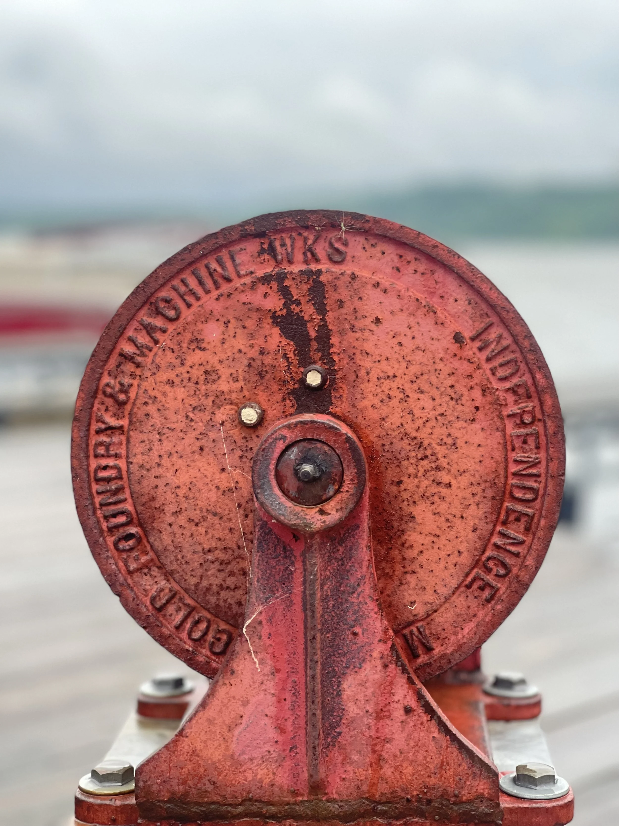 Close-up of a red, rusted fire hydrant valve with a circular top and bolts around the base, with a blurred outdoor background.