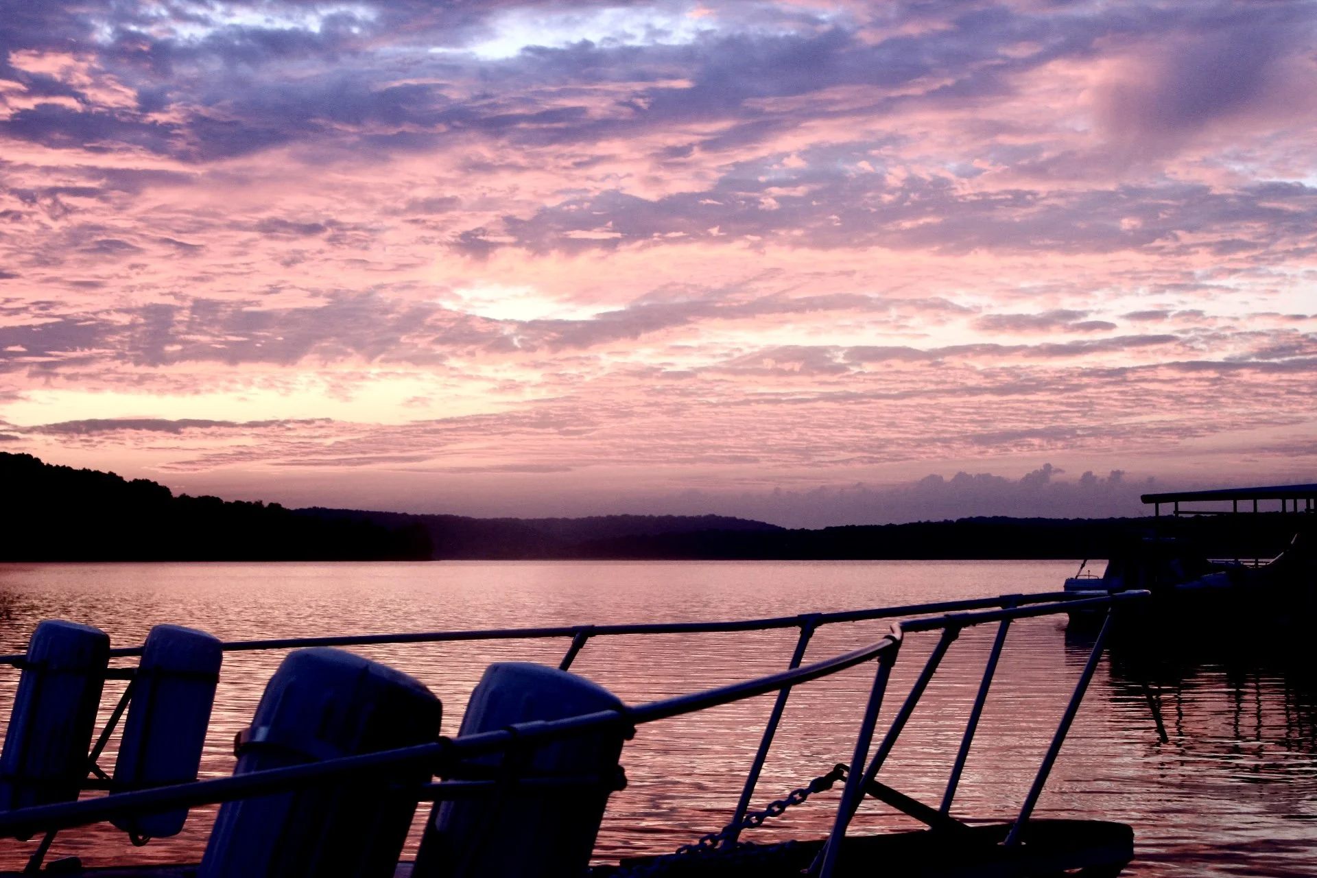 Sunset over a lake with pink and purple clouds, a boat dock and chairs on the dock in the foreground.