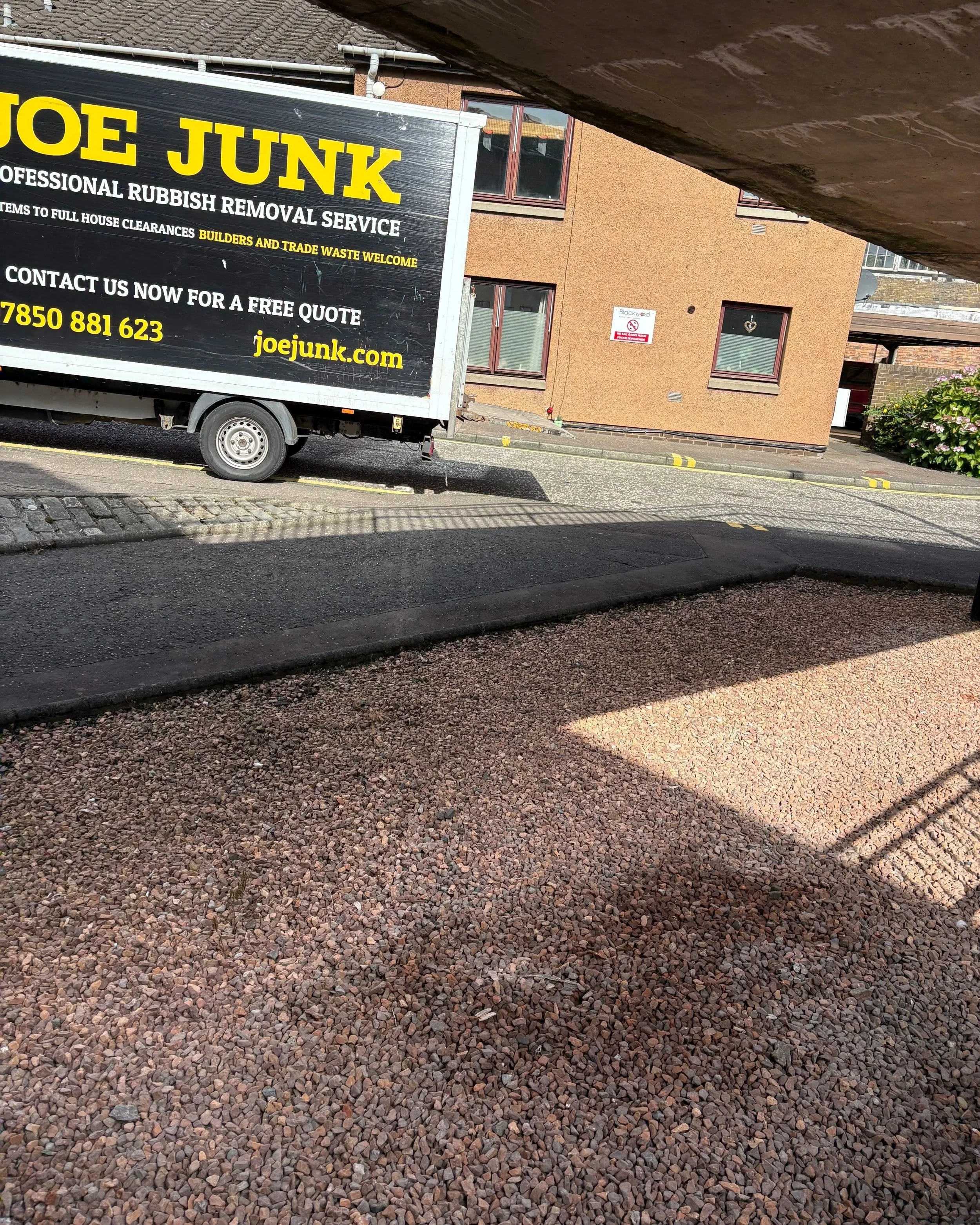 A black truck with yellow and white text advertising Joe Junk and rubbish removal services, parked on a residential street with a pebble driveway and a brown multi-story building in the background.
