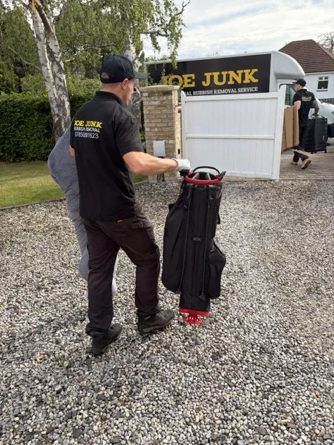 A worker from Joe Junk trash removal service moving a golf bag on a gravel driveway, with a white trash removal trailer in the background.