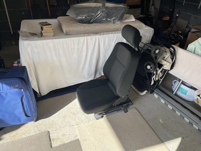 A black car seat with headrest placed on a floor next to a table covered with a beige cloth, with items like a pillow and plastic-covered mattress on top. There is a blue bag to the left and various objects on the right.