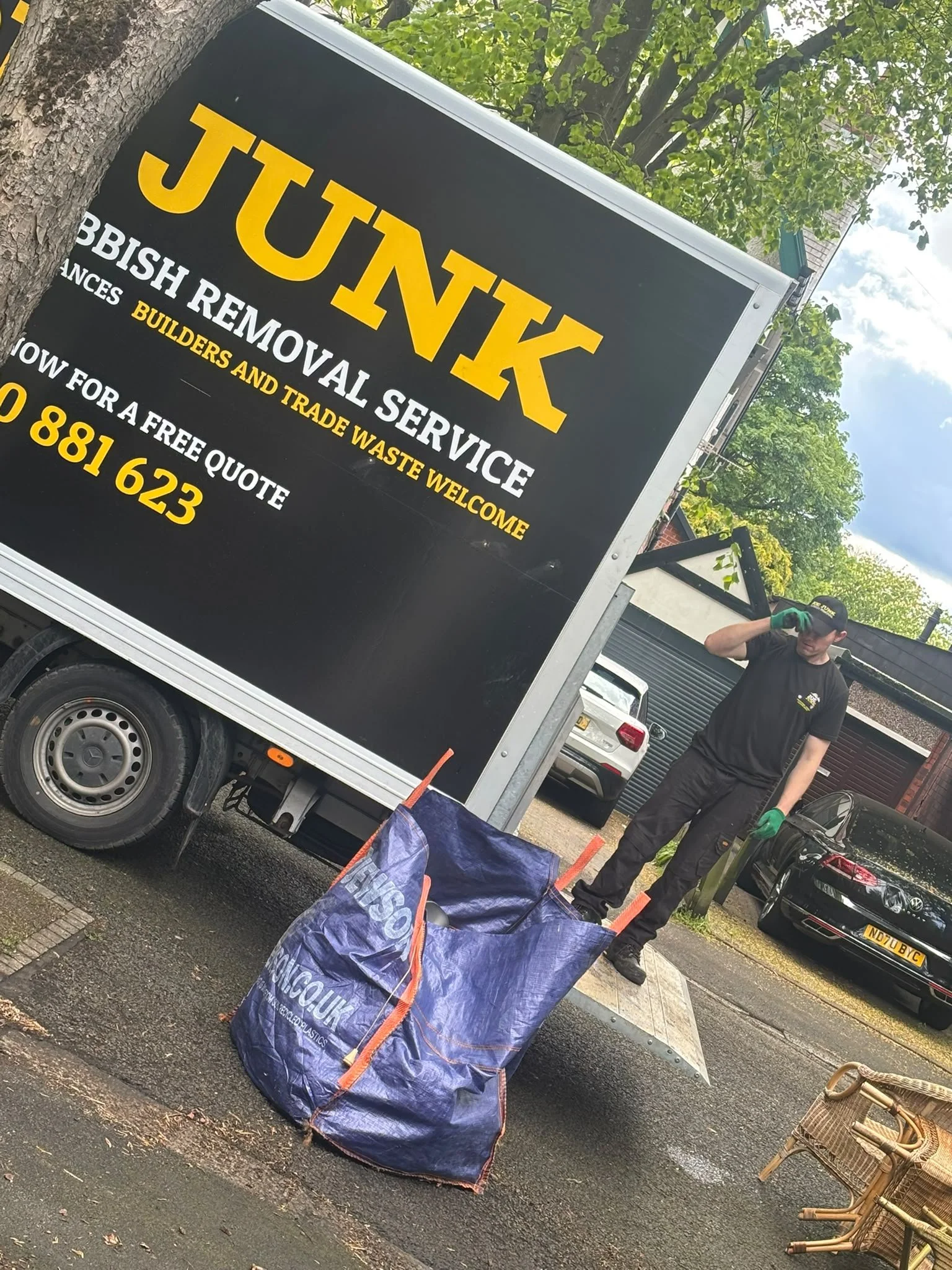 Black and yellow sign for a rubbish removal service, with contact information. In the background, a person wearing a black uniform and green gloves standing next to a large blue bag, other cars parked on the street, a tree, and outdoor chairs.