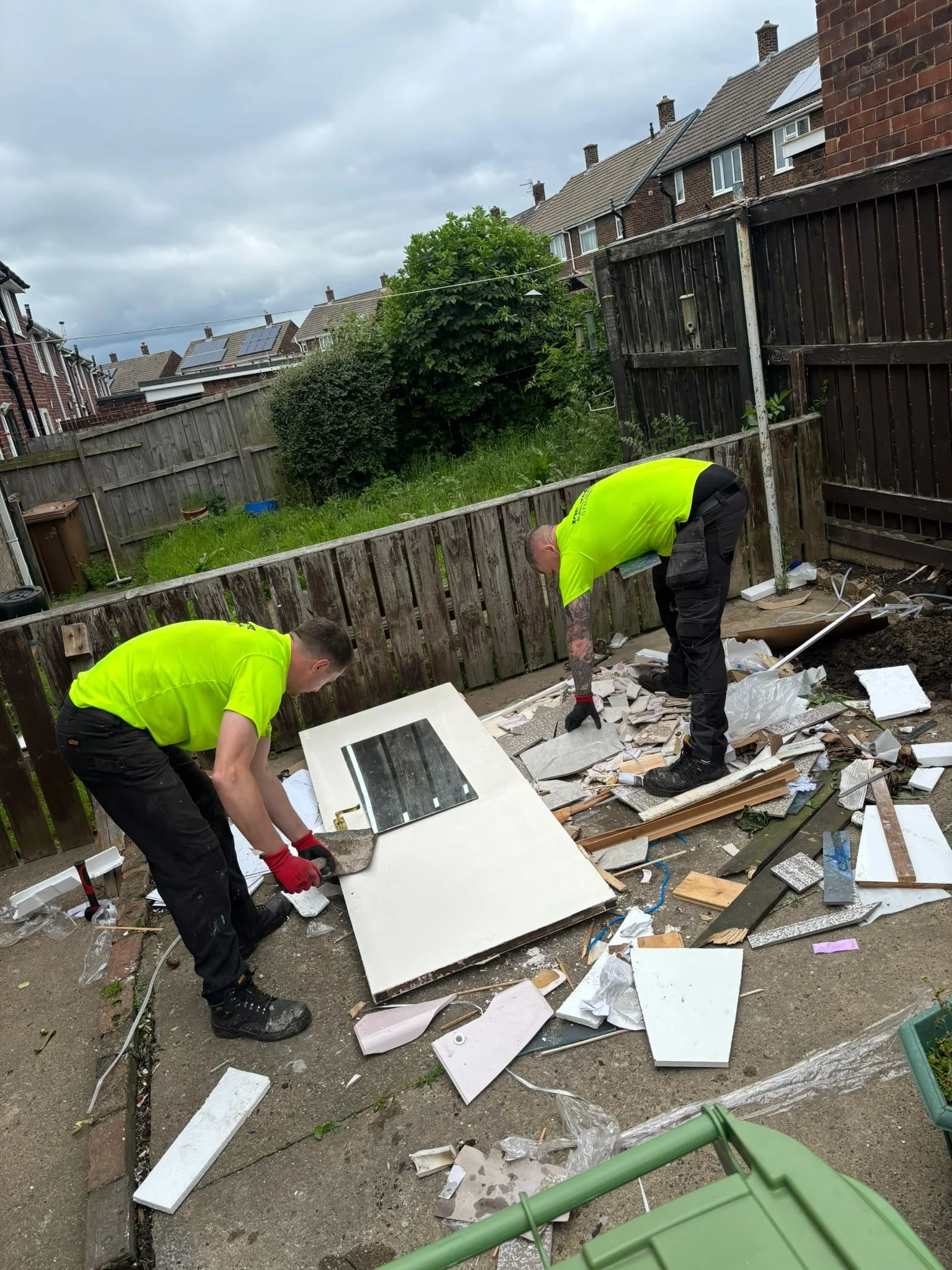 Two workers in yellow shirts and black pants are breaking apart white kitchen cabinets outside, surrounded by debris, in a backyard with a wooden fence, green grass, and neighboring brick houses.