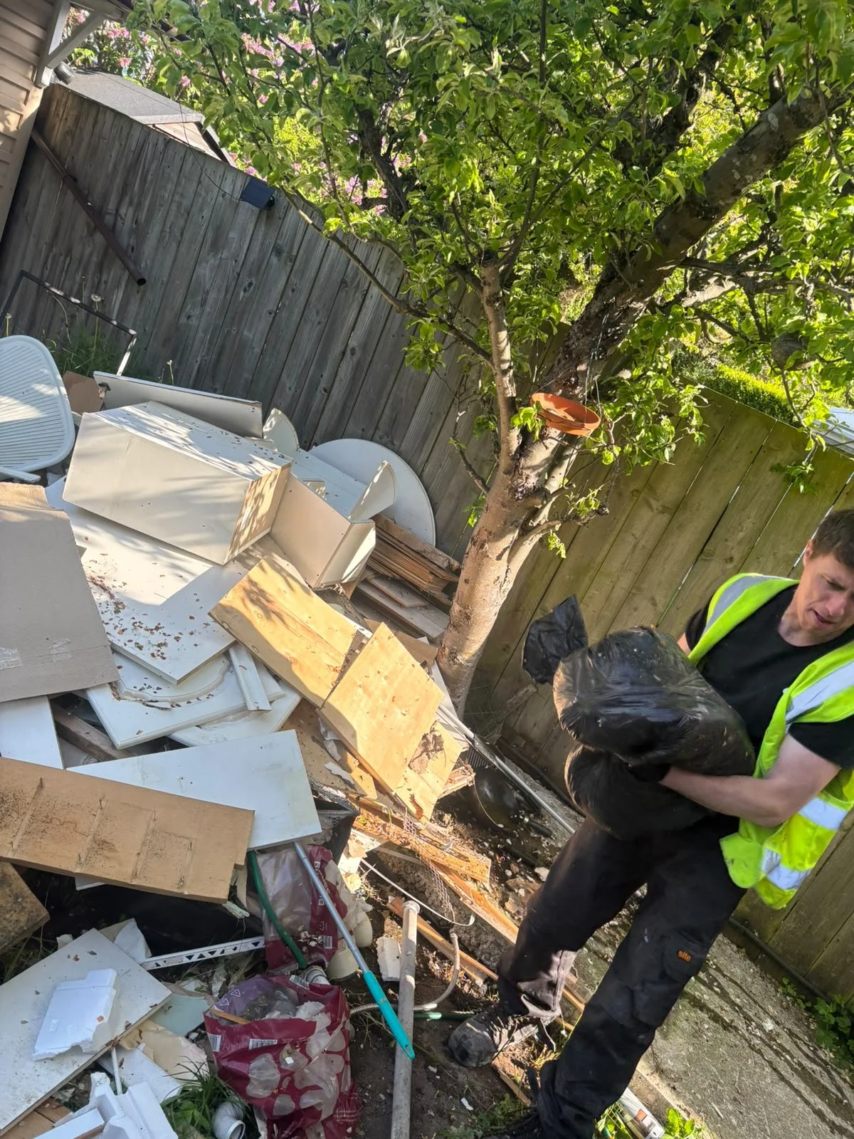 A man wearing a black shirt and a yellow safety vest holding a black trash bag at a cluttered outdoor space with debris, broken furniture, wood scraps, and a tree with a tree branch and a small orange container hanging on it.