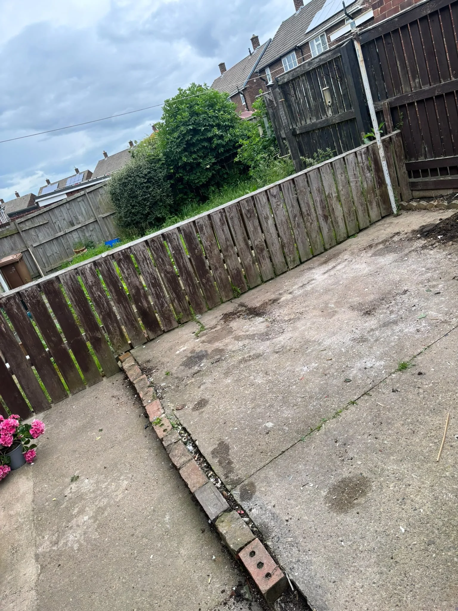 A backyard with a wooden fence, some bushes, and a concrete patio. There are houses with chimneys in the background.