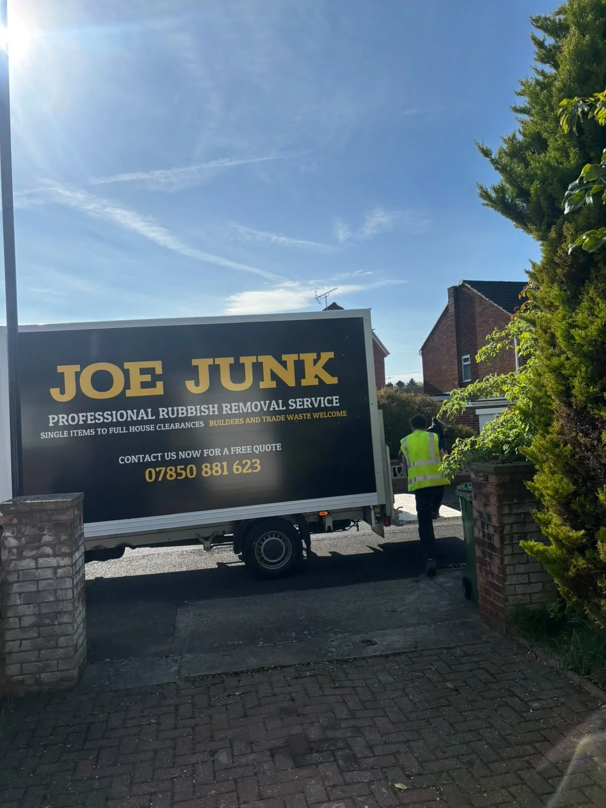 A truck with a sign advertising Joe Junk, a professional rubbish removal service, parked on a residential street with a person in a yellow safety vest walking away from the truck.
