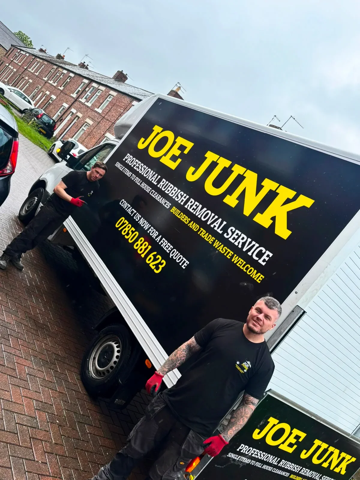 A man standing next to a black truck advertising Joe Junk, a rubbish removal service, with contact information and description of services, on a paved outdoor area with parked cars and brick buildings in the background.