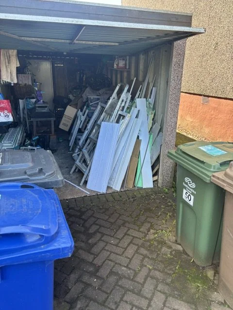 A cluttered outdoor shed filled with various household items, including white panels, a ladder, and miscellaneous objects, with two large trash bins in front on a paved surface.