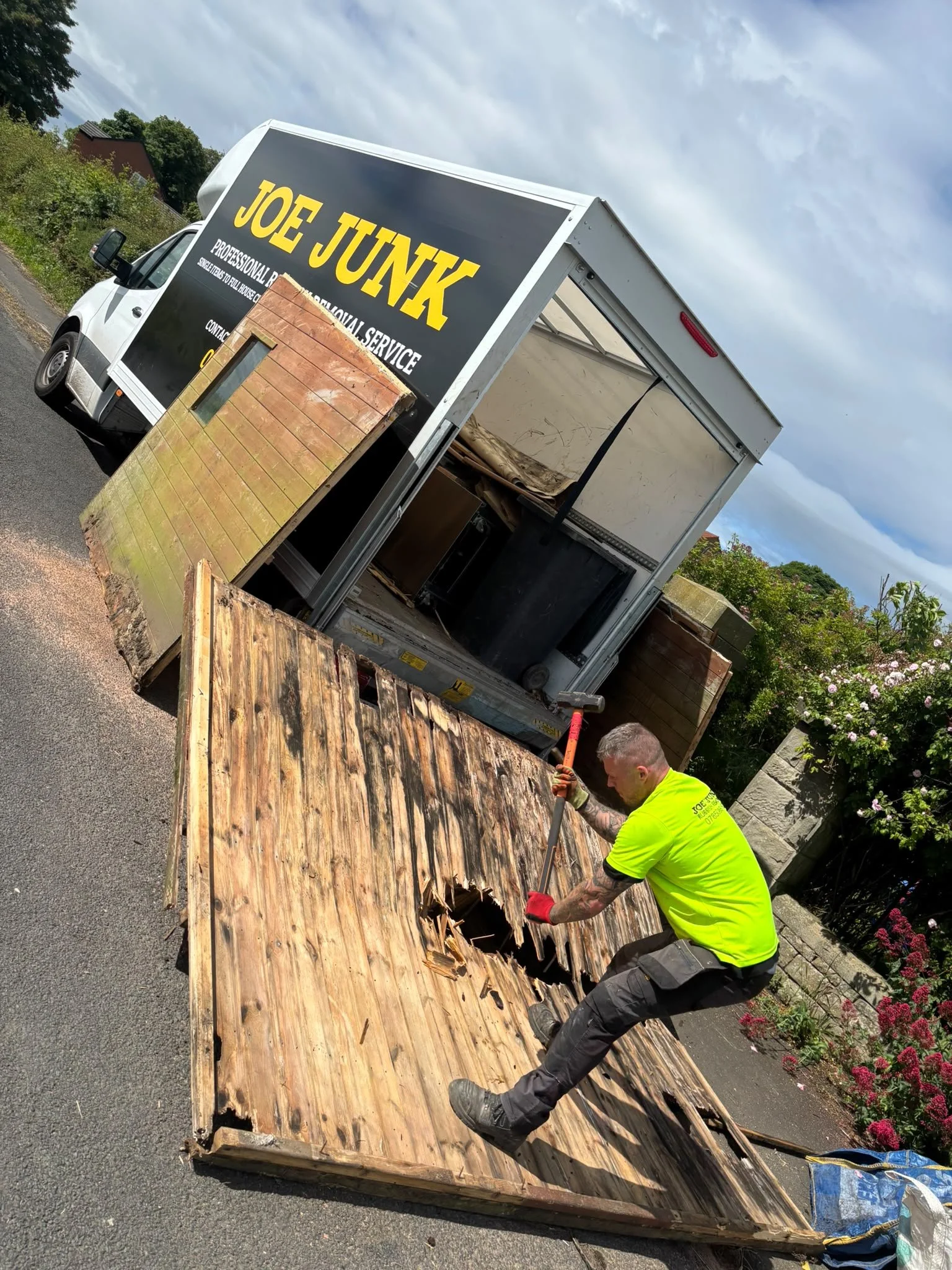 Worker in a neon yellow shirt removing damaged wooden panels from a large wooden structure, with a moving truck labeled 'Joe Junk' parked nearby, on a sunny day with partly cloudy skies and flowering bushes in the background.