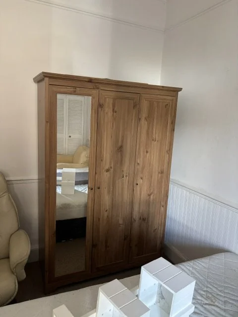 Wooden wardrobe with a mirror on the left sliding door, situated between a beige armchair and a bed in a bedroom.