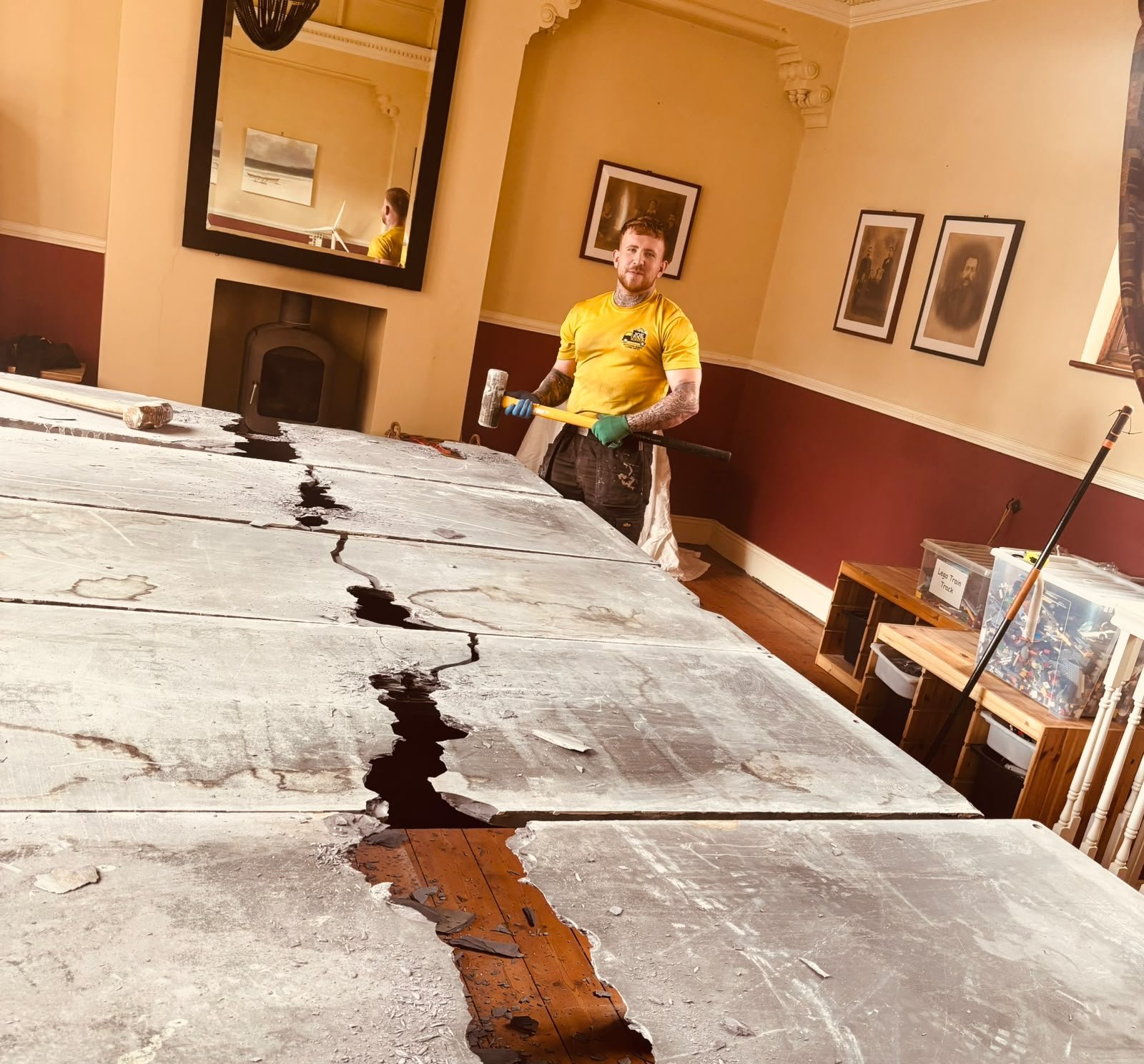 A man in a yellow shirt and gloves holding a hammer, standing behind a long table covered with large cracked pieces of abstract art or sculpted material, in a decorated room with framed pictures on the walls and plastic storage boxes in the background.