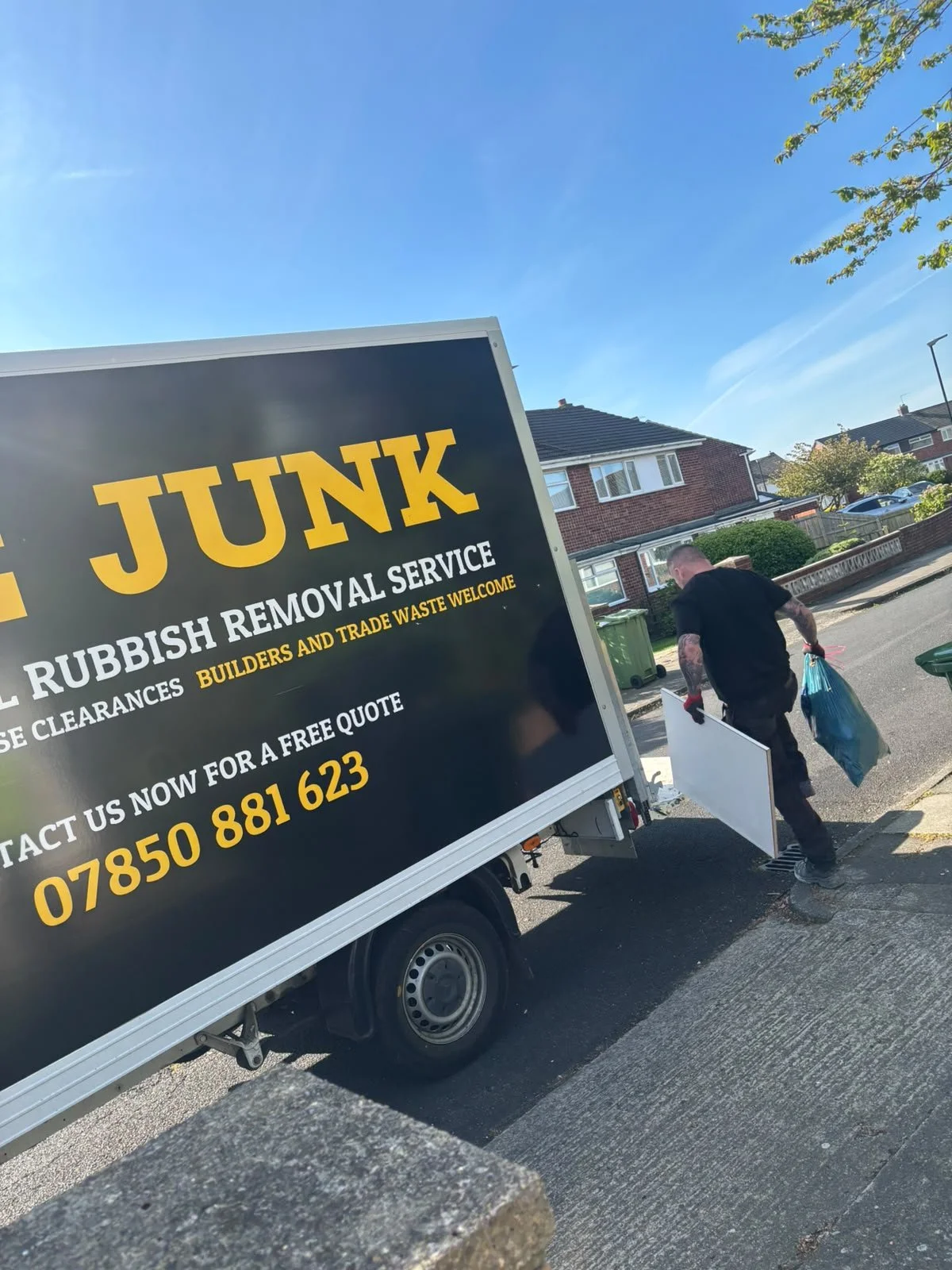 A man loading waste bags into a rubbish removal truck on a residential street. The truck has an advertisement for a rubbish removal service with a phone number.