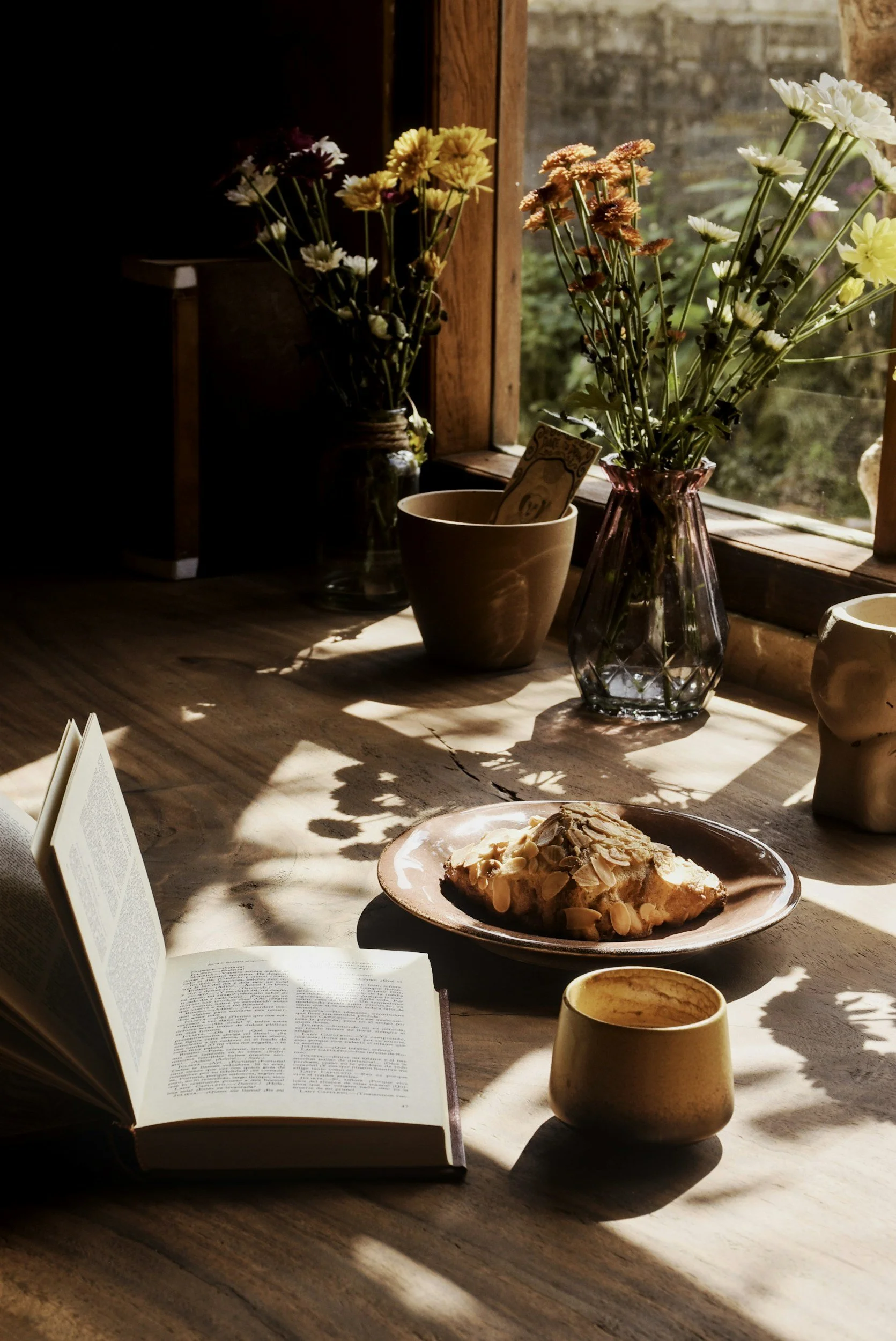 Sunlit wooden table with books, ceramic cup and fresh flowers, creating a calm reading and seasonal living atmosphere.