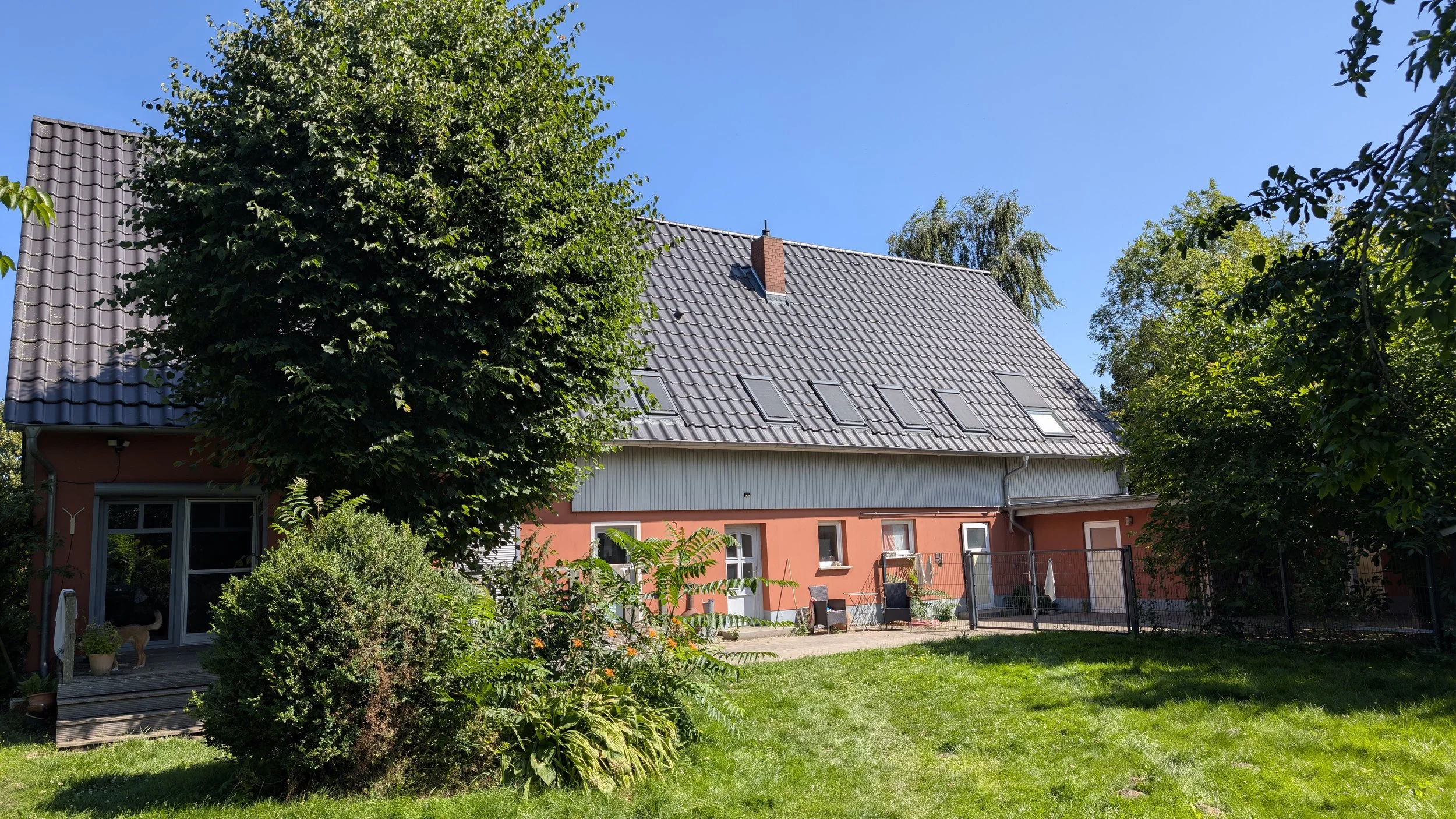 Ein Haus mit rotem Unterbau, grauem Satteldach und mehreren Dachfenstern, umgeben von Bäumen und einem Garten mit Rasen, Pflanzen und einem kleinen Balkon.