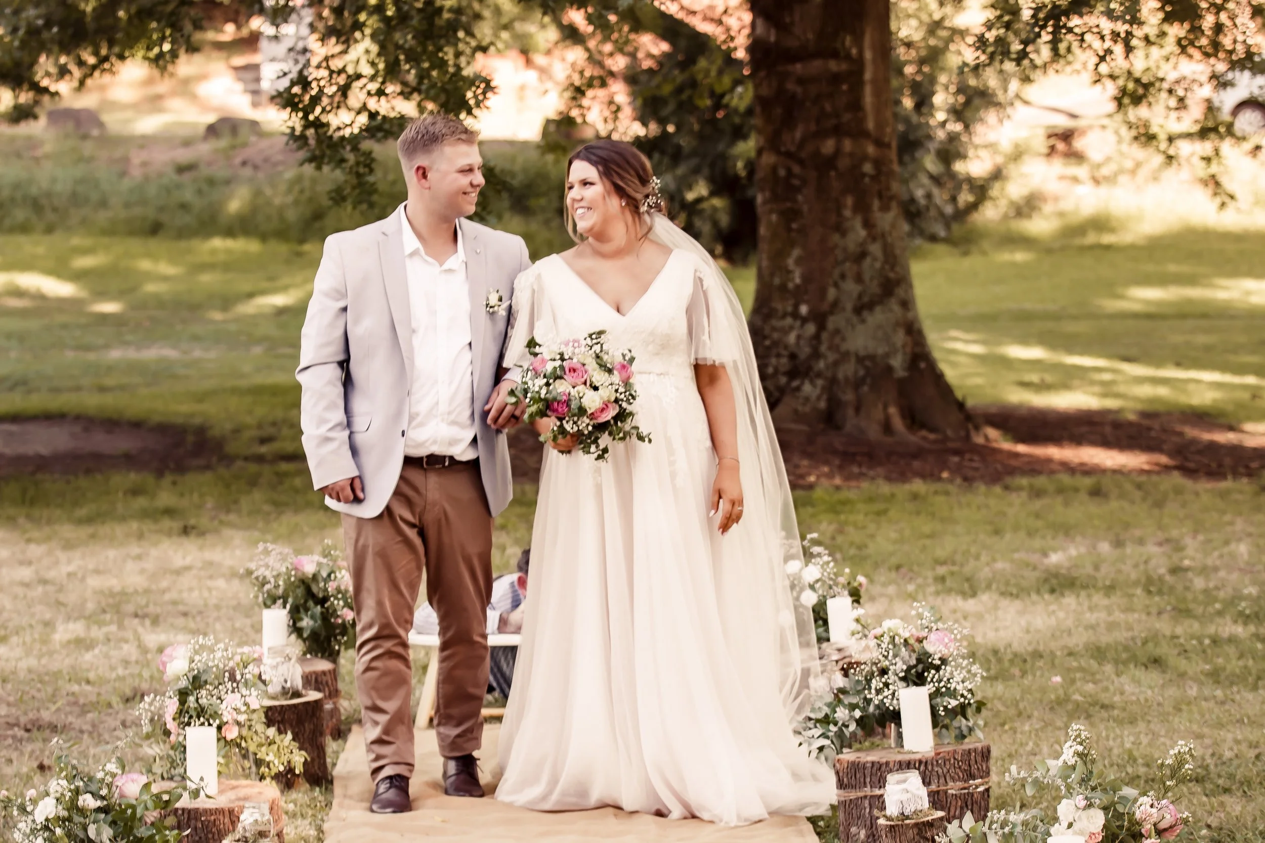 A smiling bride and groom walking together outdoors during a wedding ceremony in a park with trees and sunlight filtering through the leaves.