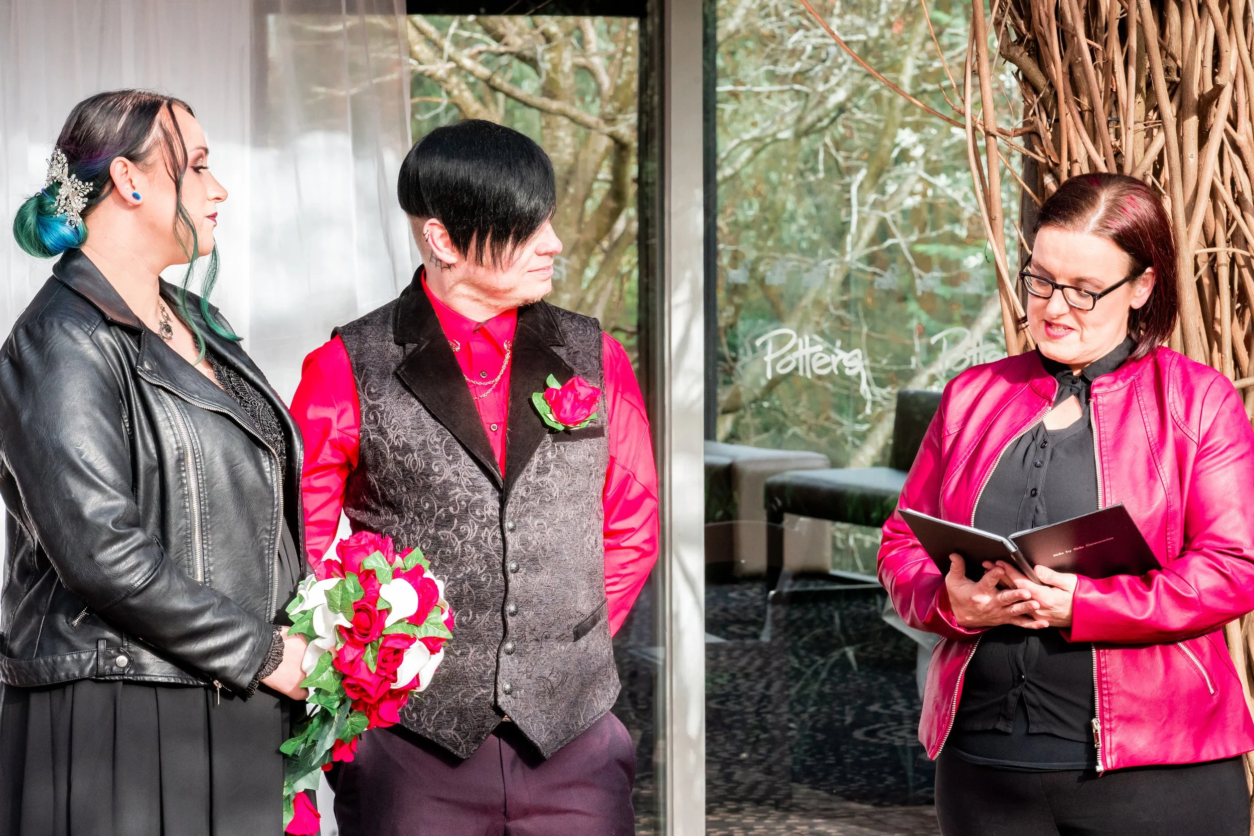 Three people standing together indoors near a window. A woman on the left holds a bouquet of pink and white flowers with greenery. The man in the middle wears a red shirt with a black vest and has a flower pinned to his vest. The woman on the right wears glasses and a red jacket, reading from a small booklet.