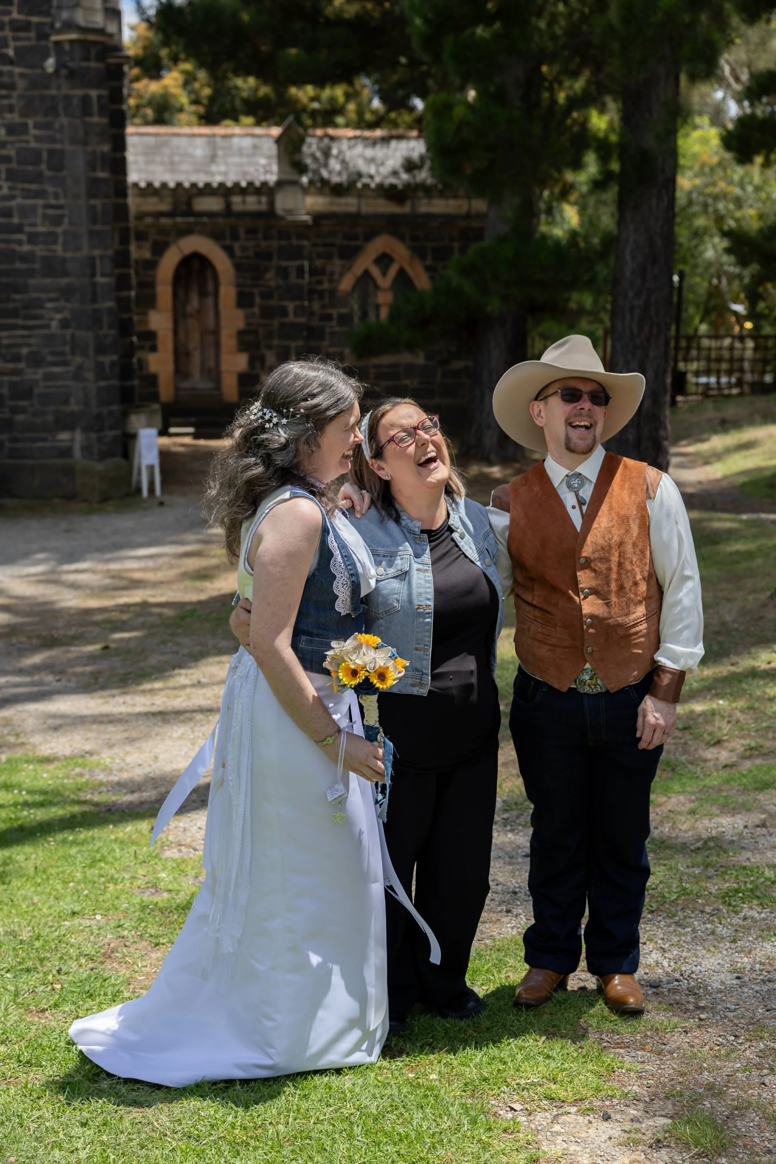 People smiling and laughing outside at a wedding, with a woman in a white dress holding a bouquet of flowers, a woman in glasses wearing a denim vest, and a man in a cowboy hat and vest.