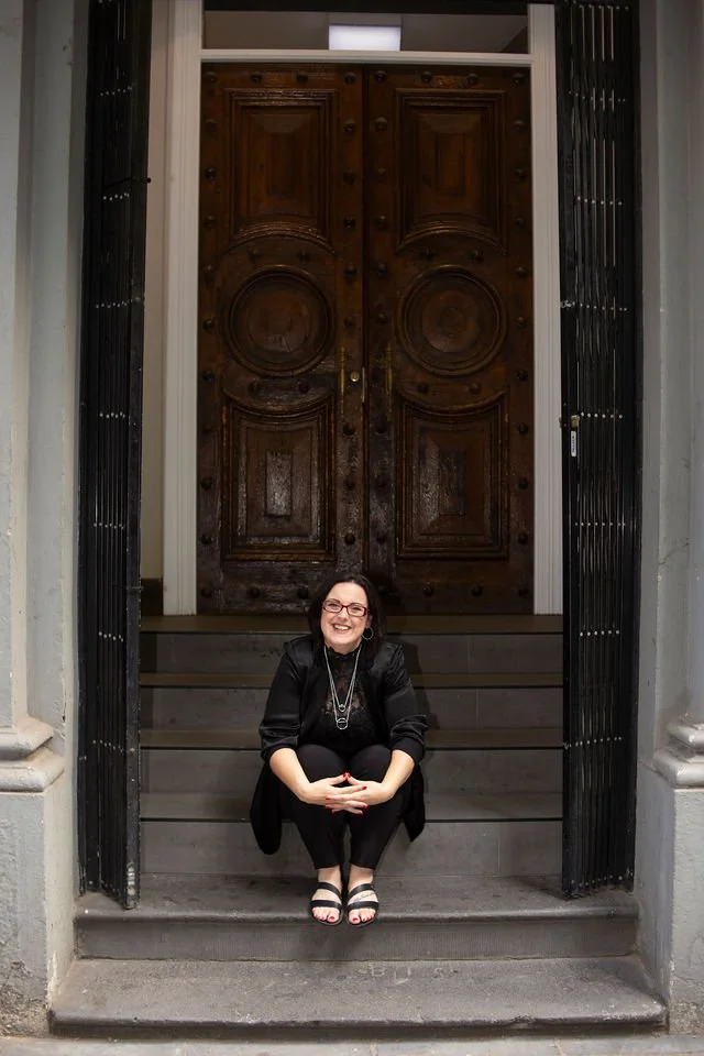 A woman with glasses and long dark hair sitting on the steps in front of a large ornate wooden door, smiling.