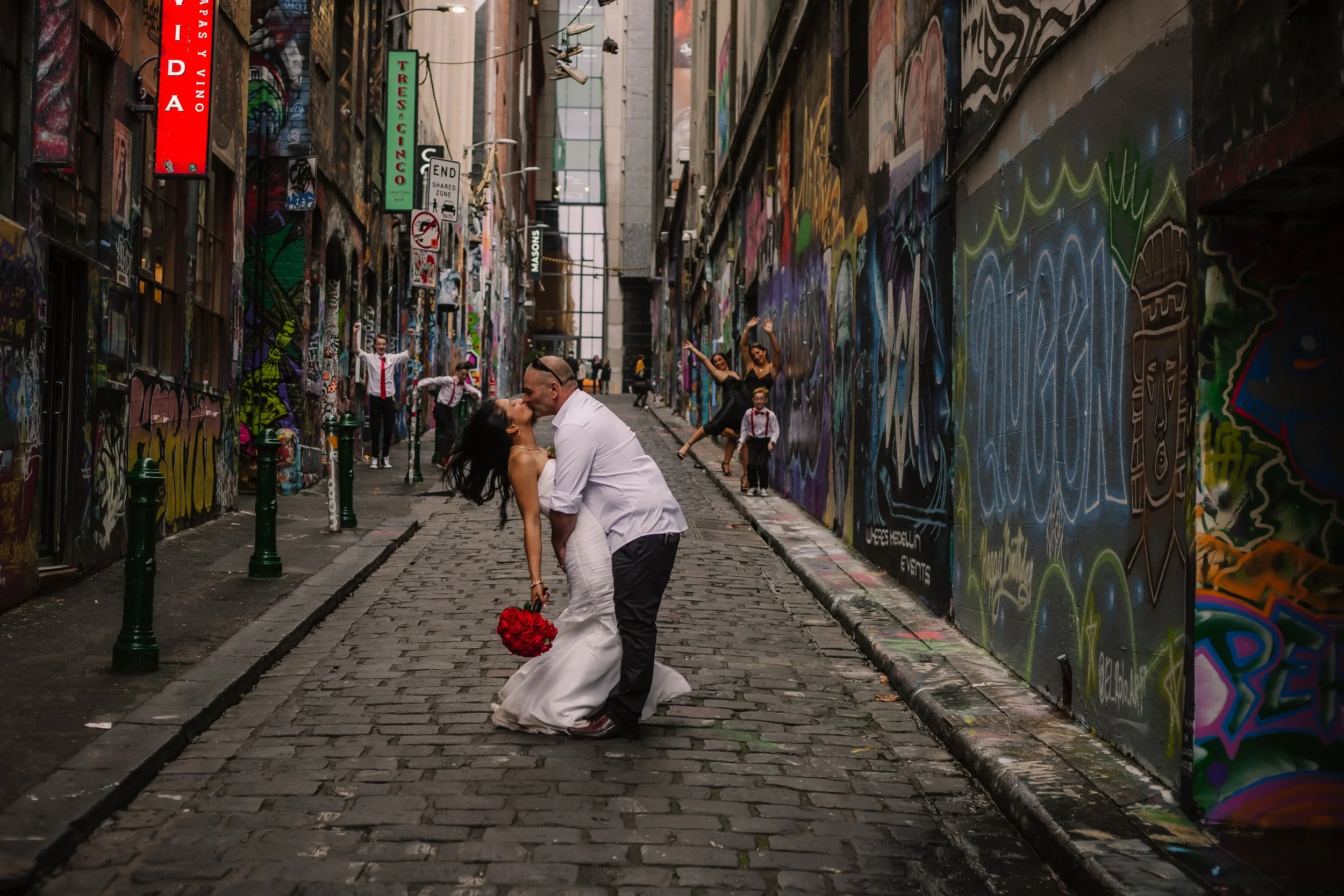 A couple dressed in wedding attire sharing a kiss in an alleyway, surrounded by colorful graffiti art with several children performing in the background.