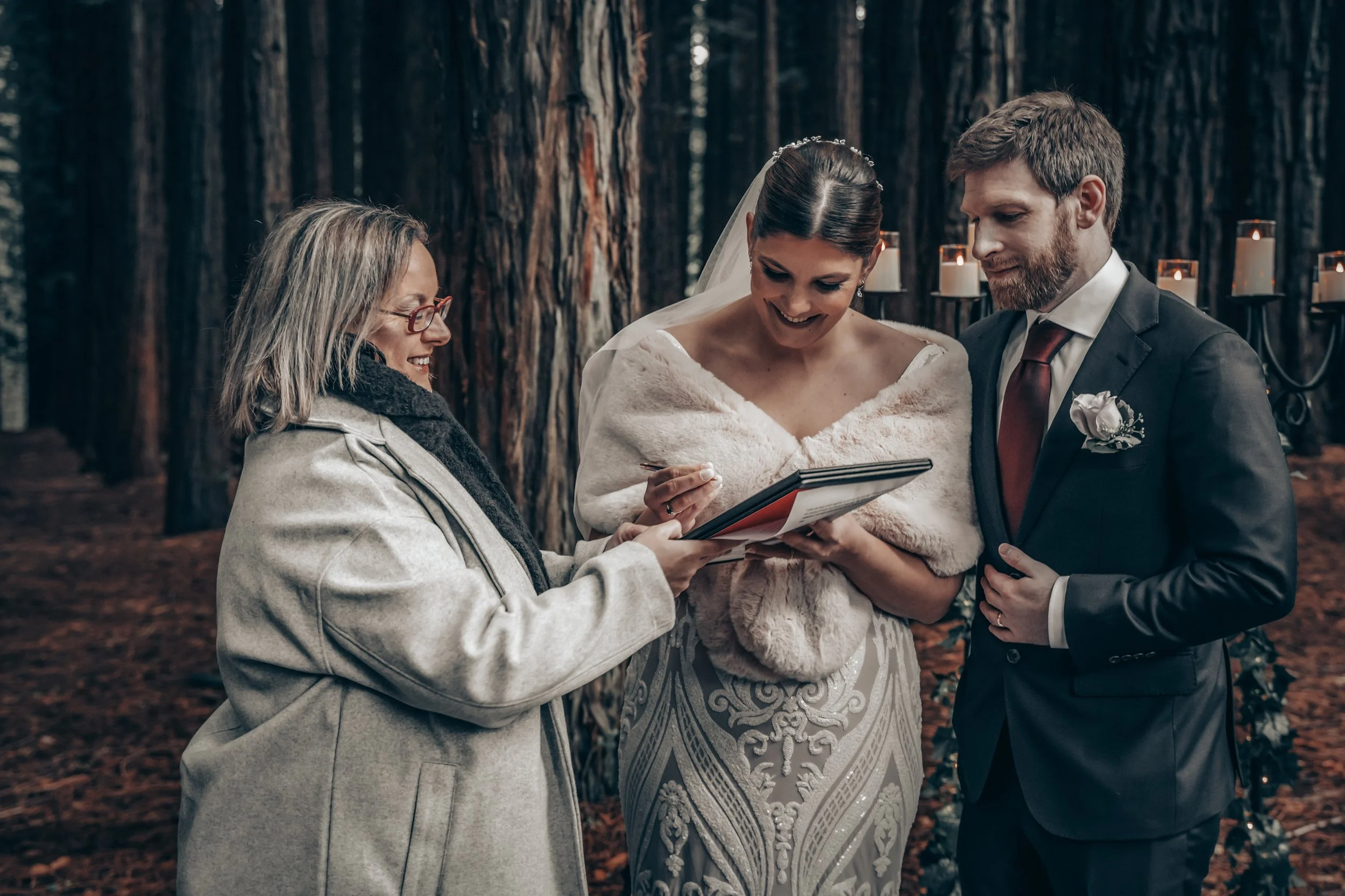 A wedding ceremony taking place outdoors in a forest, with a bride and groom exchanging vows and a woman officiant holding a book or document.