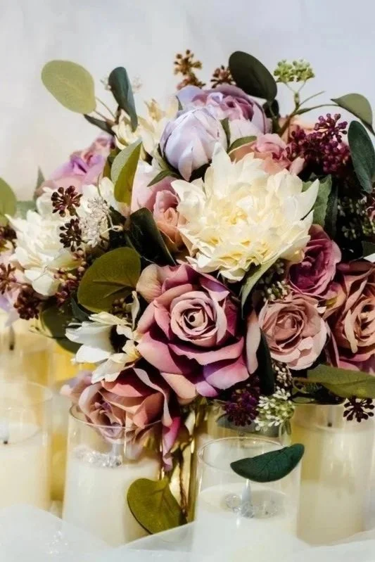 A floral centerpiece with pink, white, and purple roses, white peonies, and green foliage in three glass vases.