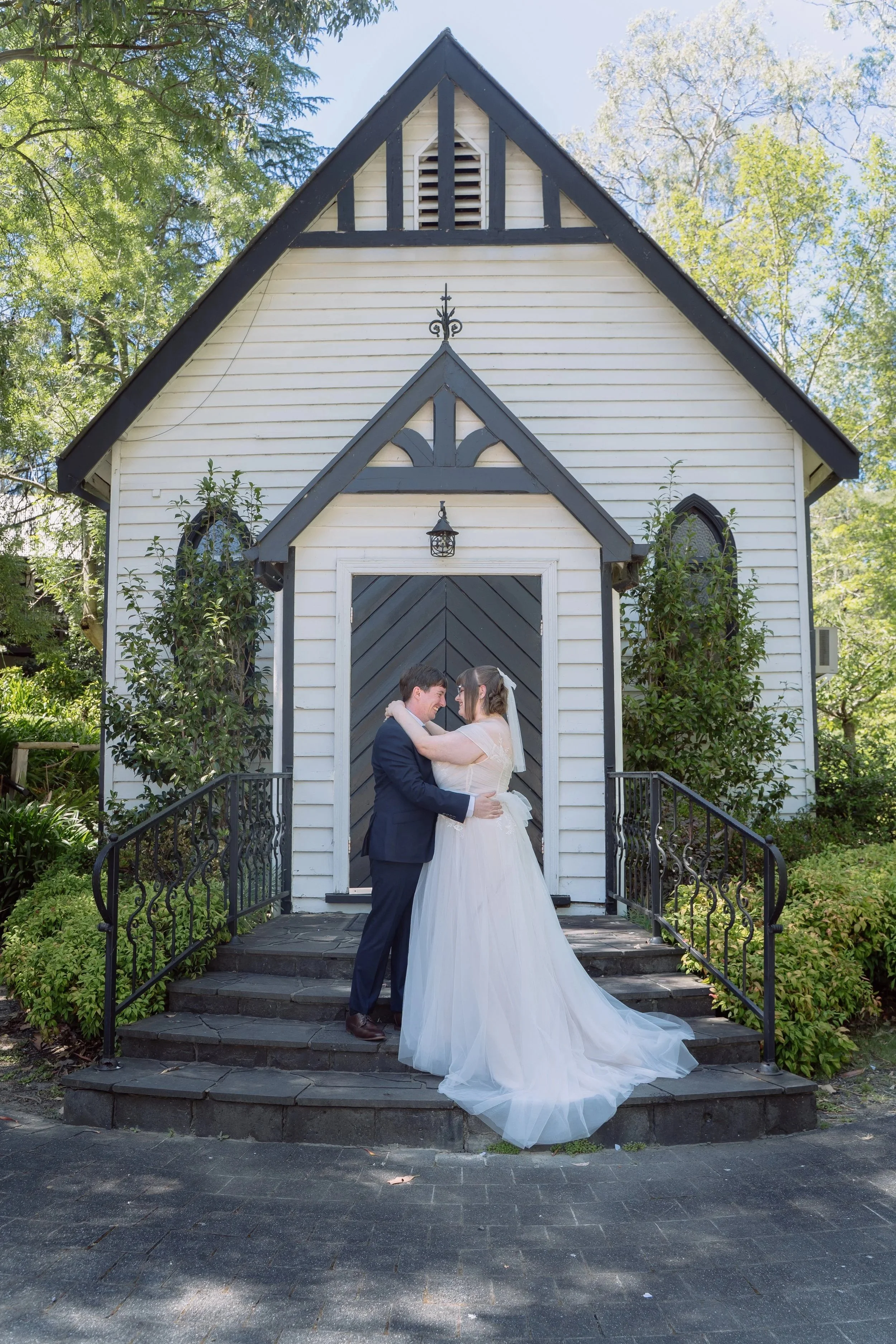 A bride and groom are embracing and smiling in front of a white wooden chapel with a dark-colored door and decorative trim. The groom is wearing a navy suit and the bride is in a white wedding gown with a long train. The chapel is surrounded by green trees and bushes.