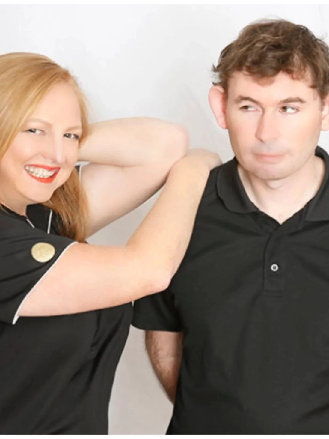 A woman with red hair and a black shirt smiling with her arm around a man with brown hair in a black shirt, both against a white background.