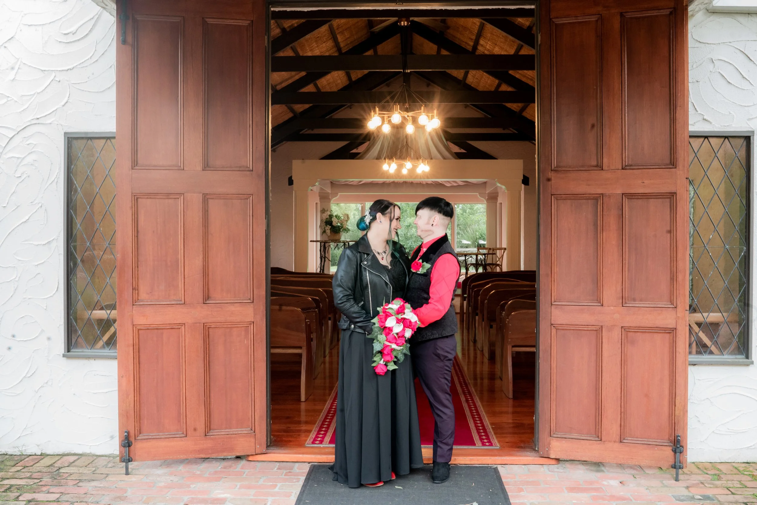 A couple standing inside a church doorway, smiling at each other, with the woman holding a bouquet of pink and white flowers, after a wedding ceremony.