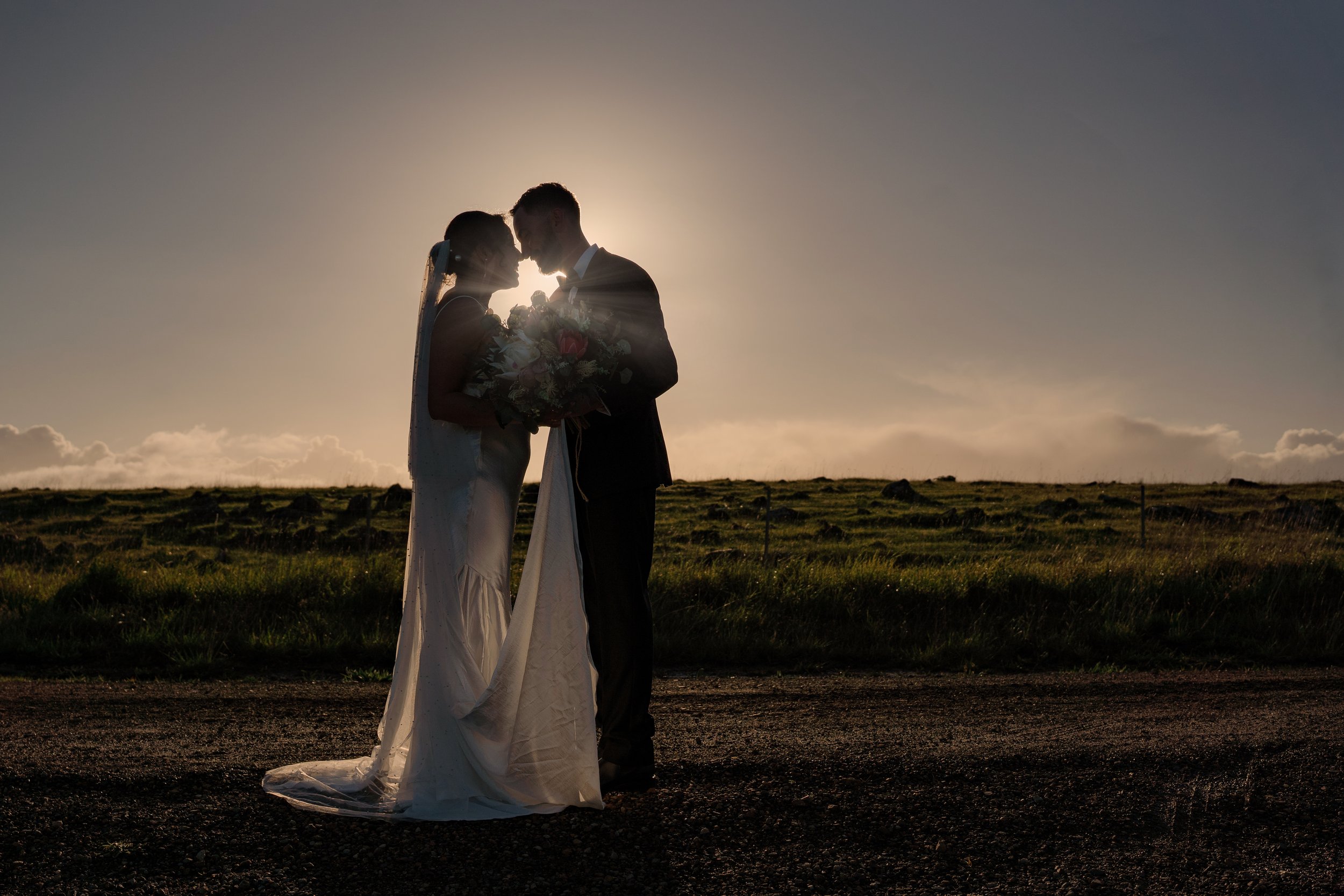 A bride and groom standing close together outdoors during sunset, facing each other with noses almost touching, holding a bouquet of flowers, with a grassy field and a partly cloudy sky in the background.