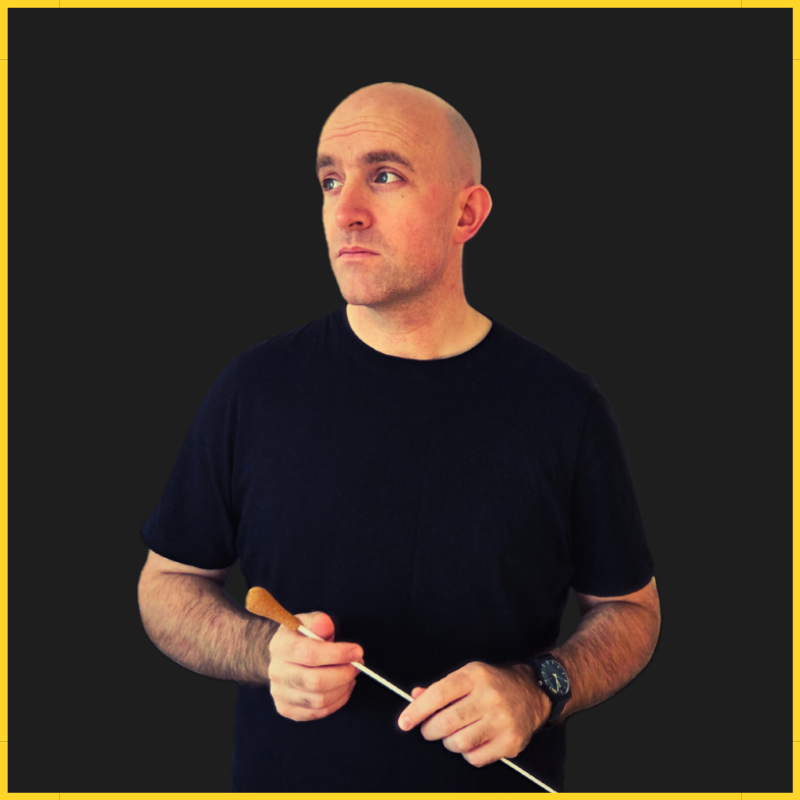 A man in a black T-shirt holding a conductor's baton against a dark background with a yellow border.