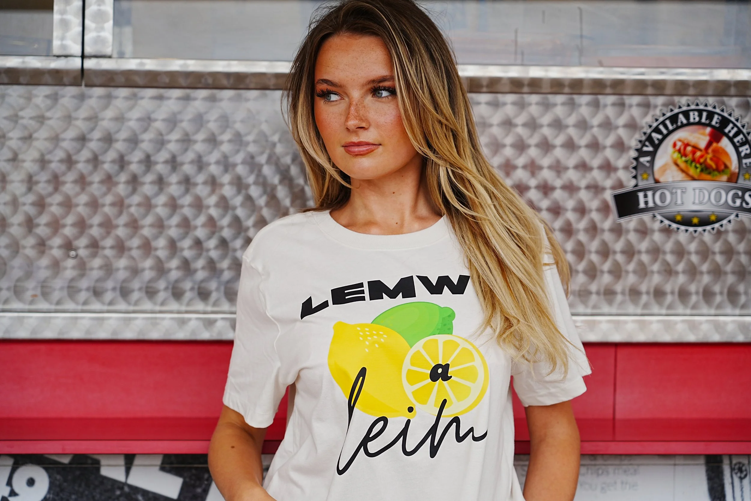 A young woman with long blonde hair, wearing a white t-shirt with lemon graphics and text, standing in front of a metallic textured background with a hot dog stand logo. This is a very cool way to show a welsh language clothing brand.