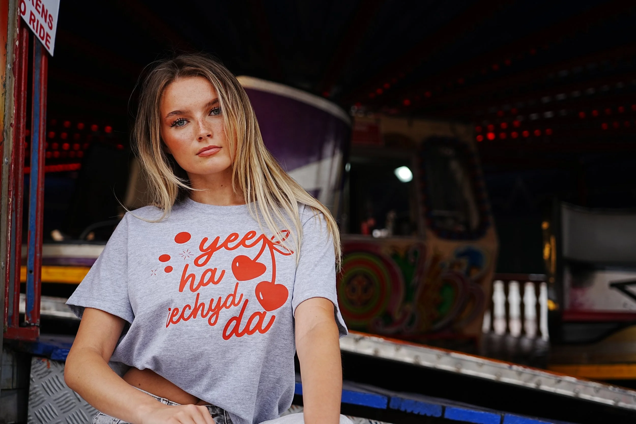 A young woman with long blonde hair and freckles standing in front of a carnival ride, wearing a gray t-shirt with red text and hearts that says 'yes ha'. This is a very cool way to show a welsh language clothing brand.
