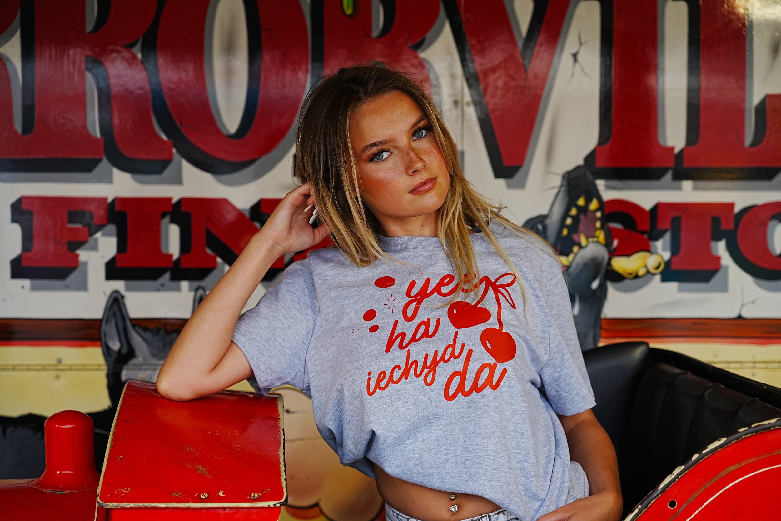 A young woman with blonde hair and blue eyes sitting on a red amusement park ride, wearing a gray T-shirt with red text and cherries, leaning her head on her hand and looking at the camera.