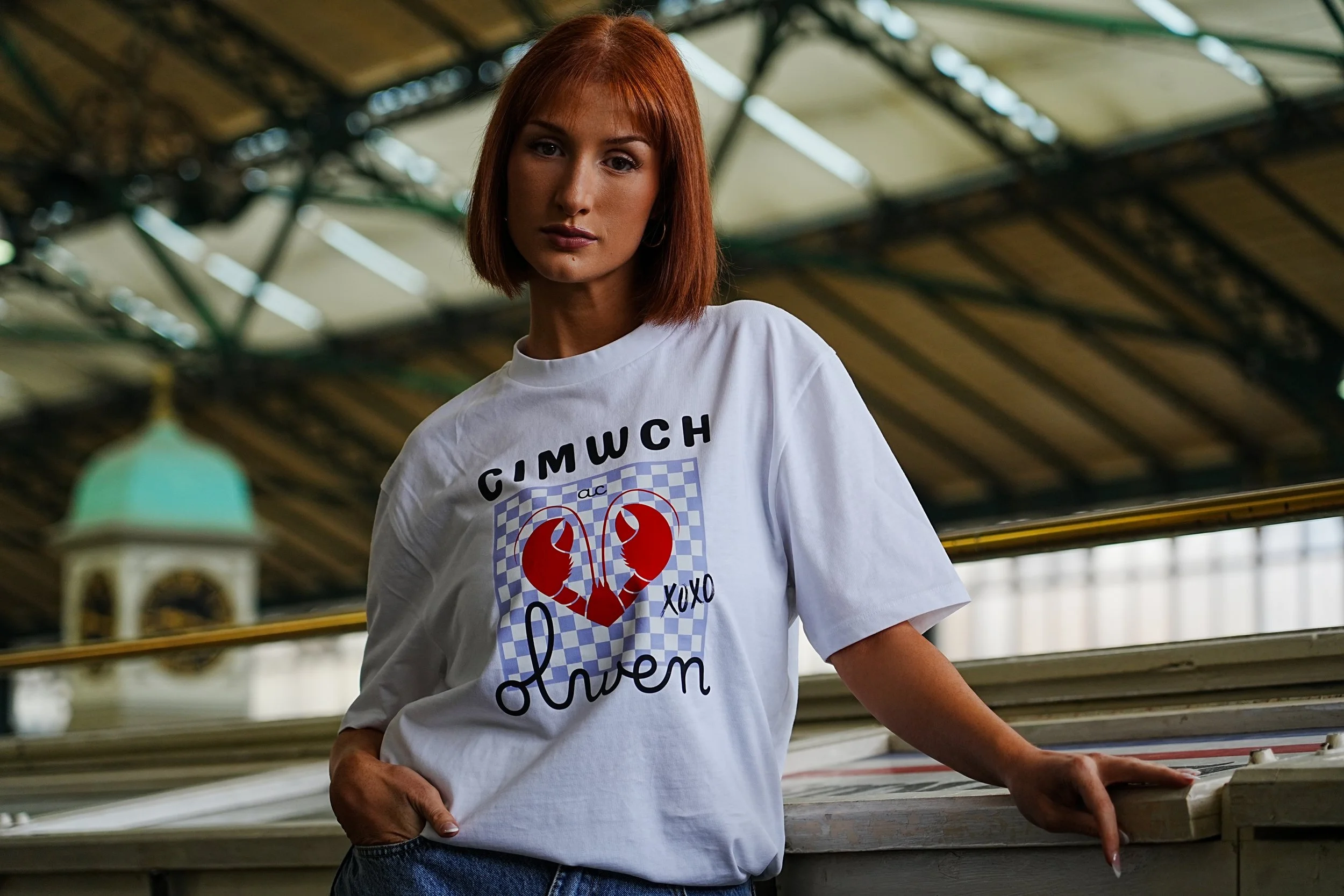 A woman with red hair wearing a white t-shirt with a lobster graphic and the words 'CIMWCH XOXO When' stands outdoors, leaning on a surface, with an ornate structure and sky in the background. This is a very cool way to show a welsh language clothing