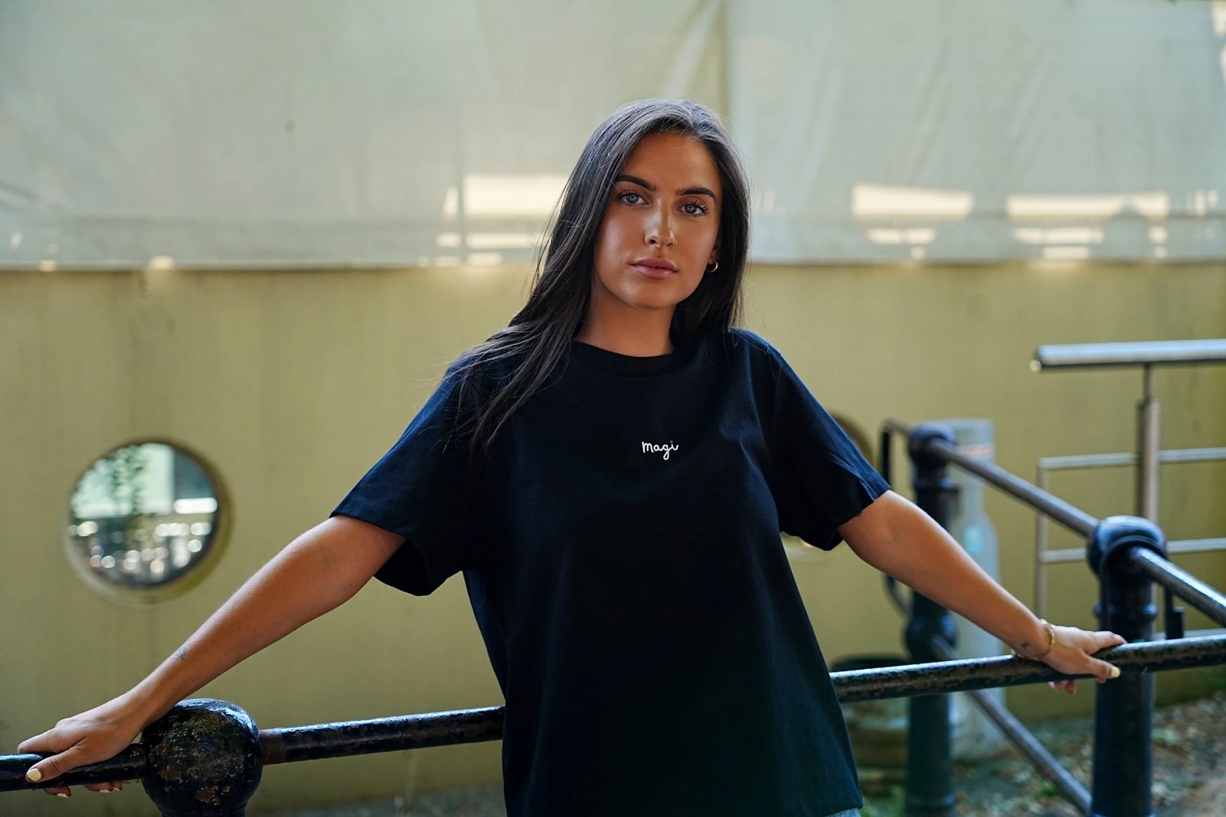 Young woman with long dark hair and light skin leaning on a railing outdoors, wearing a black T-shirt with the word 'Magi' written on it, in front of a yellow building with a circular window. This is a very cool way to show a welsh language clothing